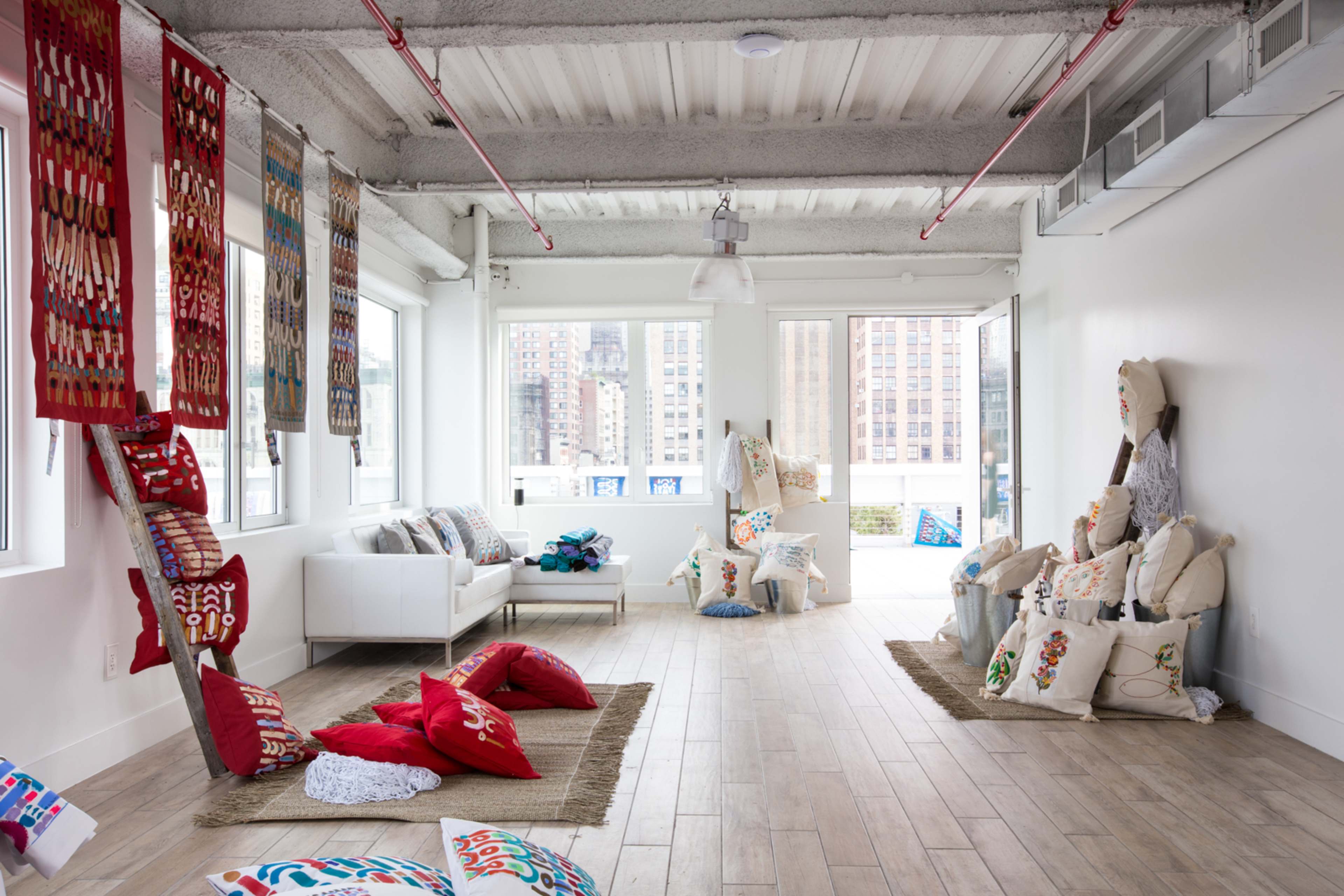 A bright, open room features a white sofa, various colorful cushions, textile art hanging on the walls, and large windows overlooking a cityscape.