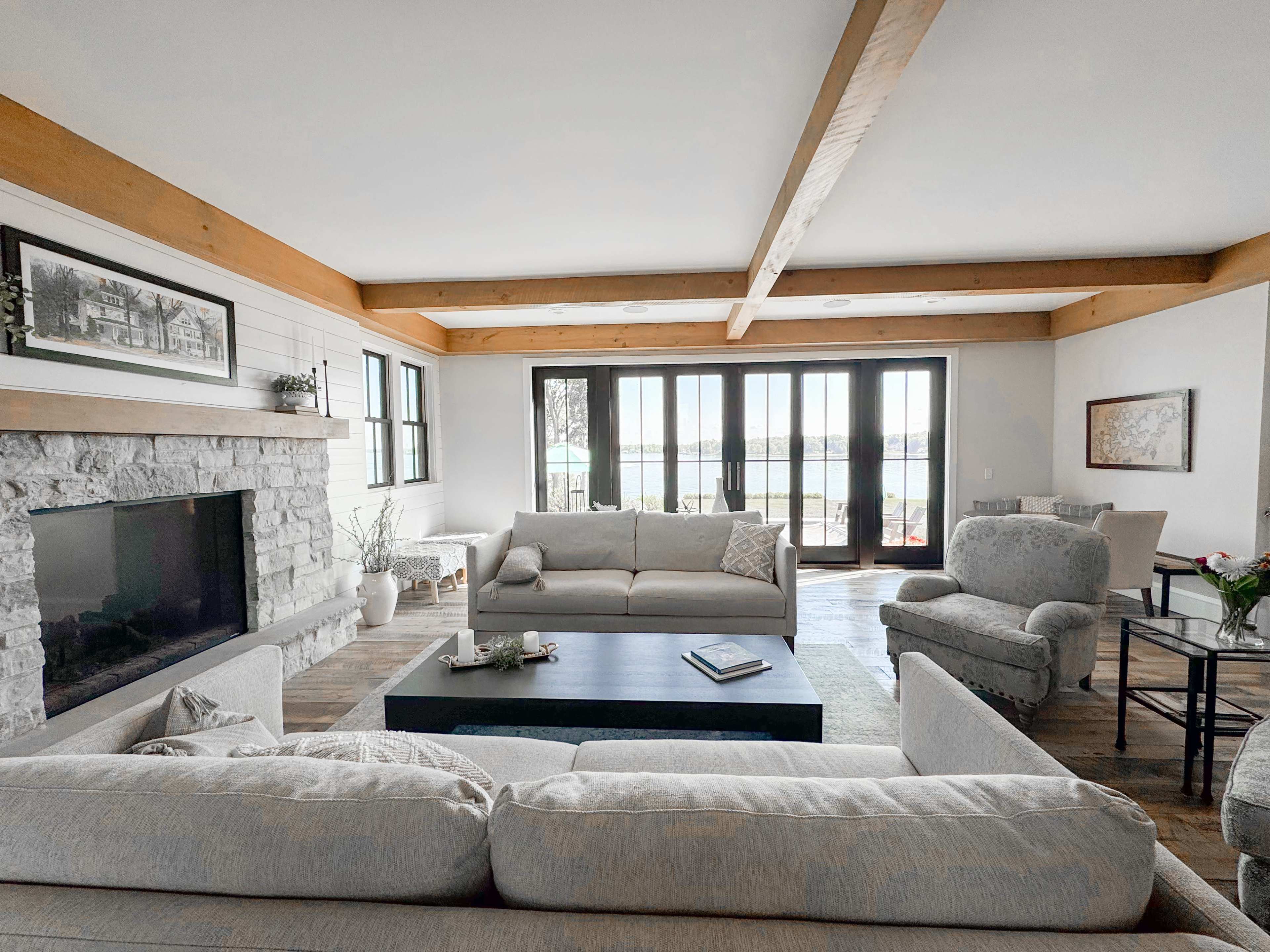 The image shows a spacious living room featuring a stone fireplace, light-colored sofas, wooden beams on the ceiling, and large windows that overlook a lake.