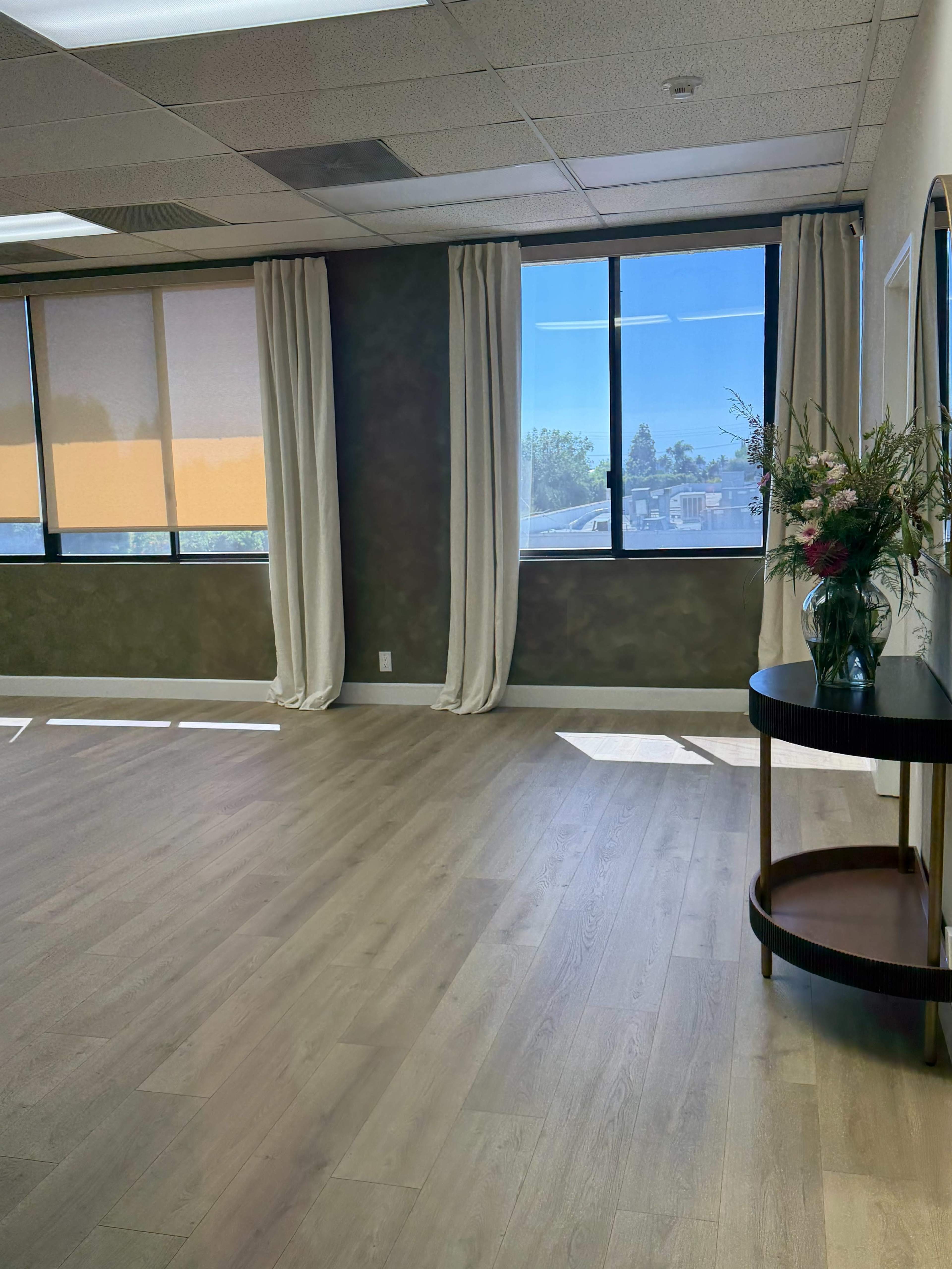The image shows a bright, spacious room with large windows, light wood flooring, a round table, and a vase of flowers on it.