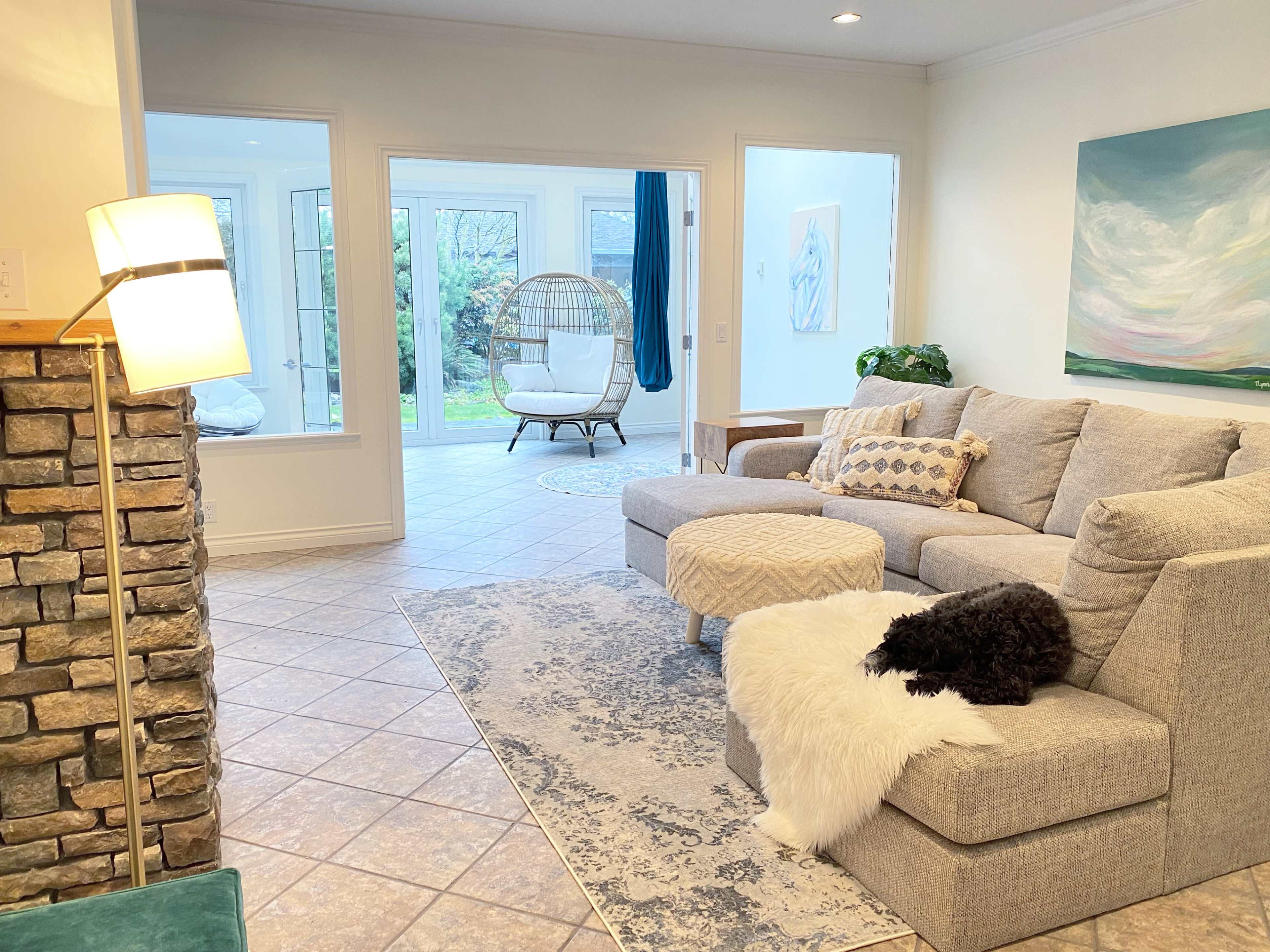 A light-filled living area with a gray sectional sofa, a stone accent wall, and a view of a sunroom featuring a hanging chair and outdoor greenery.