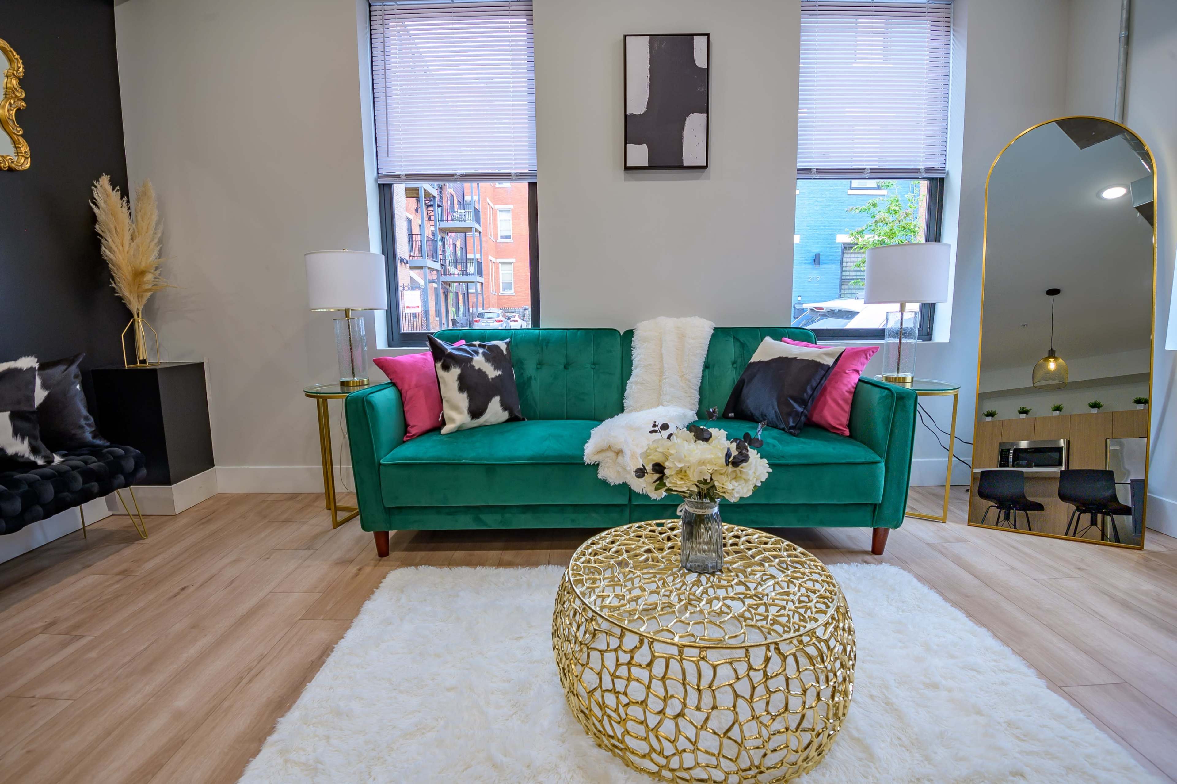 A modern living space featuring a green velvet couch with pink and black pillows, a decorative gold coffee table, and large windows allowing natural light into the room.