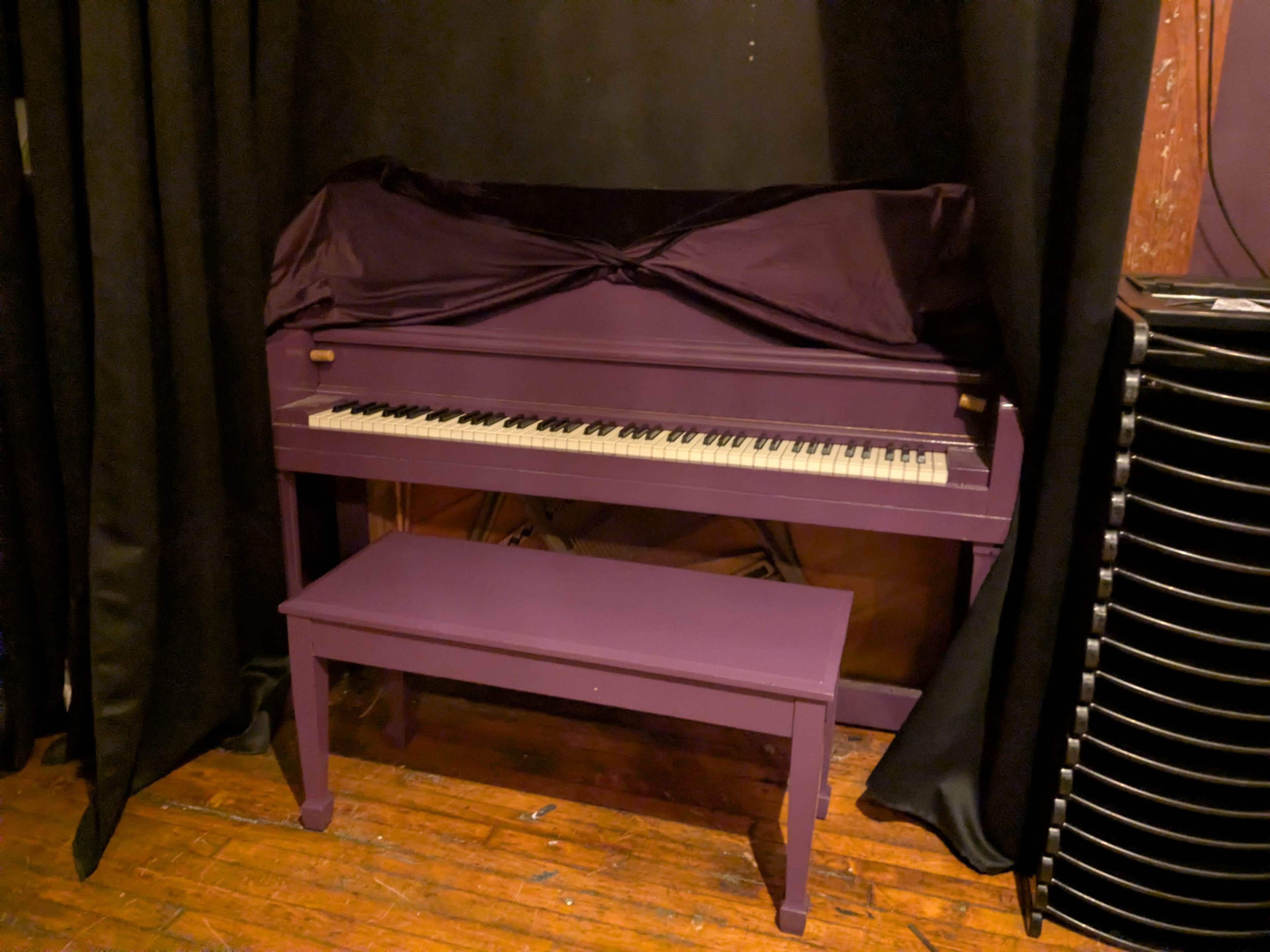 A purple piano and matching bench are positioned against a black curtain in a dimly lit space.