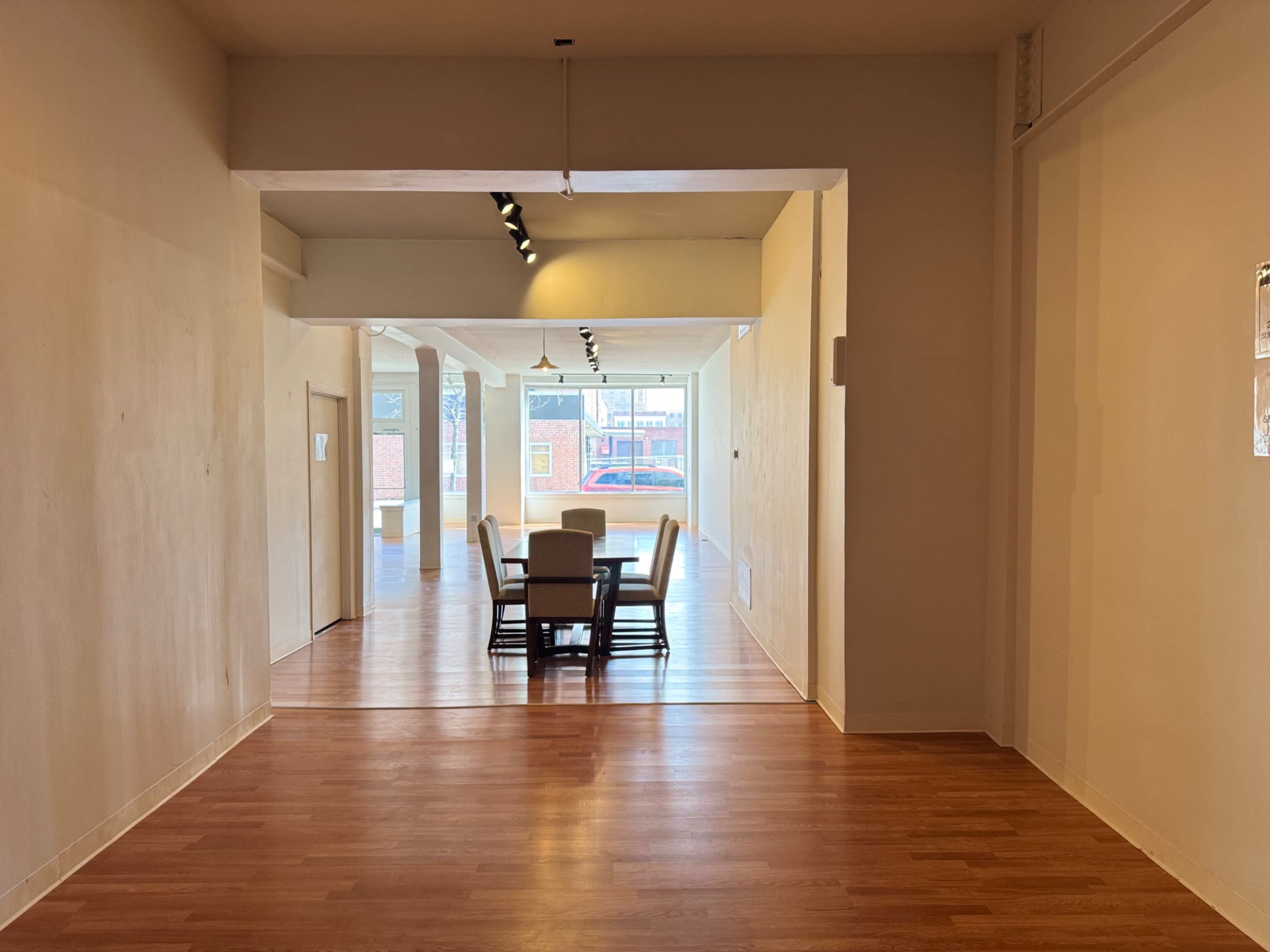 The image shows a simple interior space featuring a dining table with chairs at its center, viewed through an open passageway.