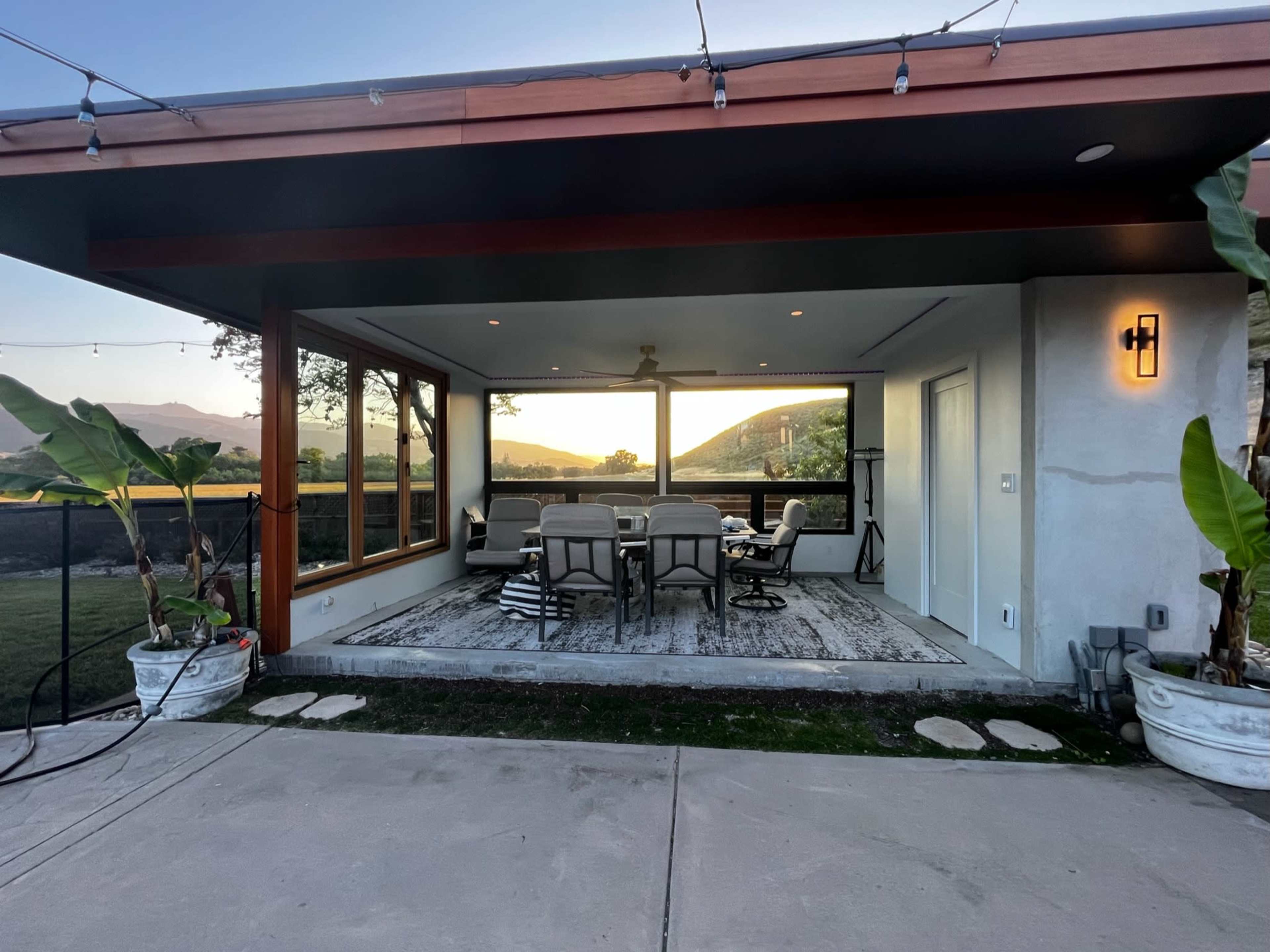 A modern covered patio features a seating area with a table and chairs, surrounded by large windows overlooking a scenic landscape.