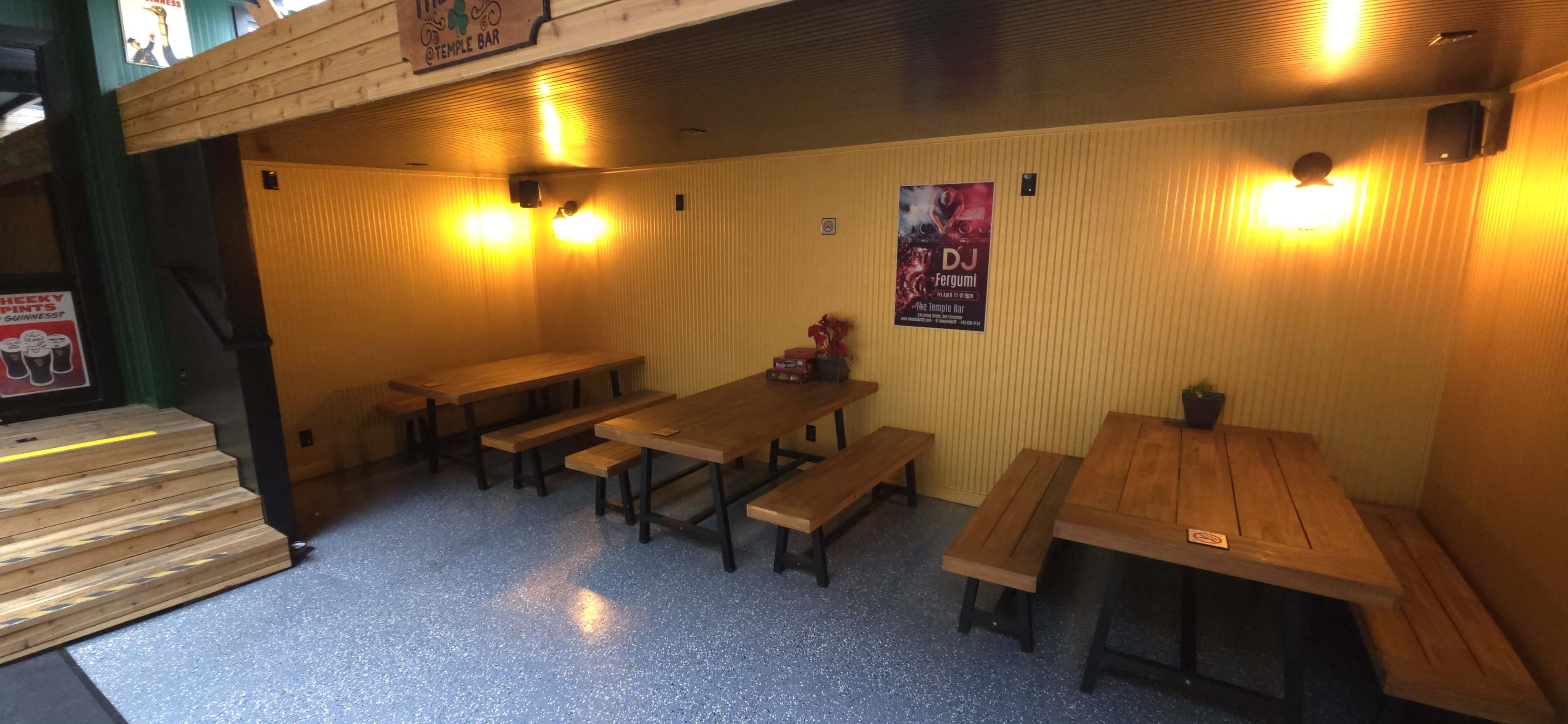 The image shows a bare, enclosed space with wooden tables and benches arranged under warm lighting, complemented by a poster on the wall and a small plant on one of the tables.