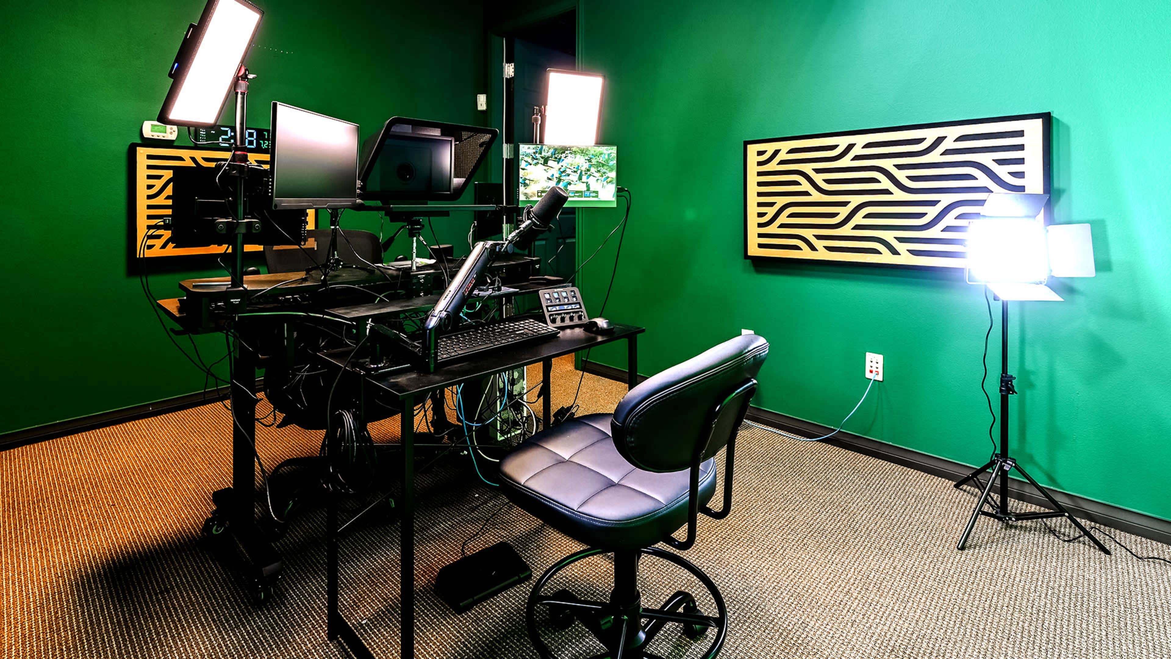 The image shows a small recording studio with a green wall, a black desk equipped with multiple monitors, a microphone, and lighting setups, alongside a swivel chair.