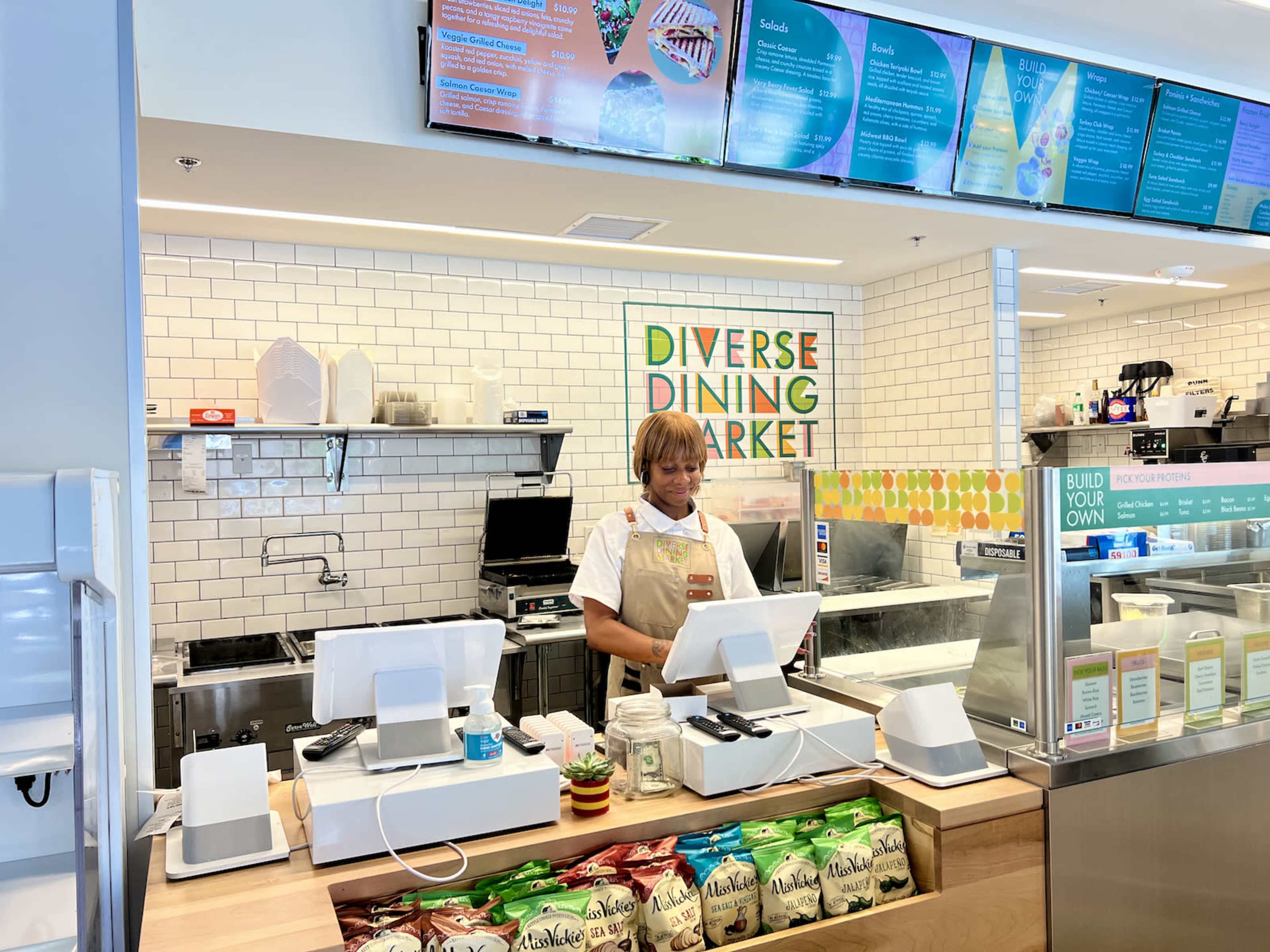 A cashier stands behind a counter at a dining market, surrounded by food displays and digital menus.