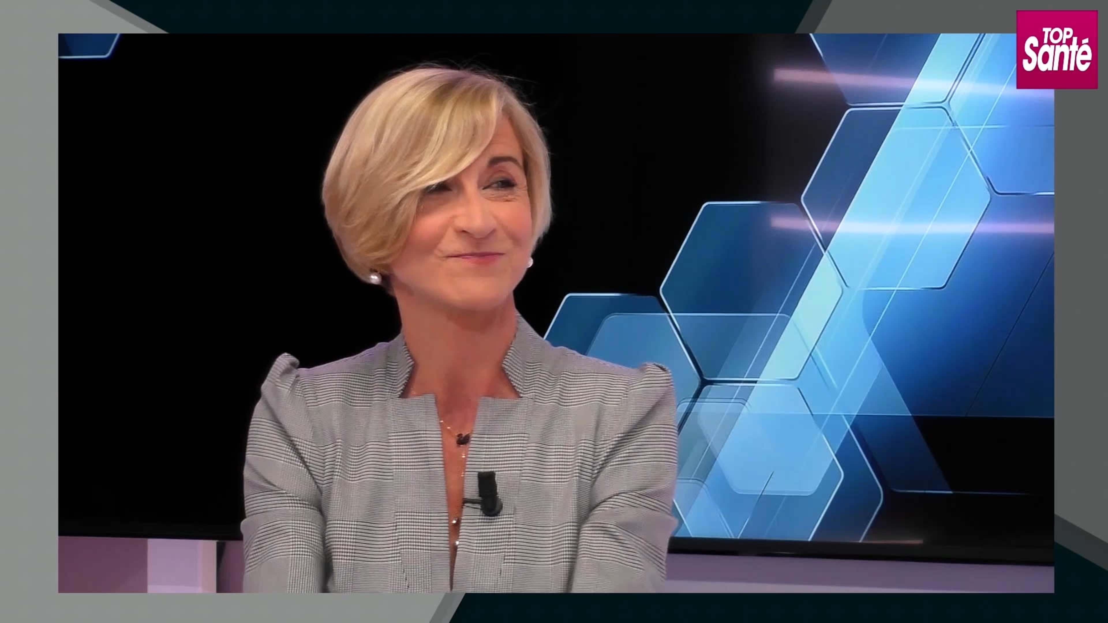 A woman with short blonde hair is sitting in front of a modern, geometric background featuring blue hexagons and a logo in the corner.