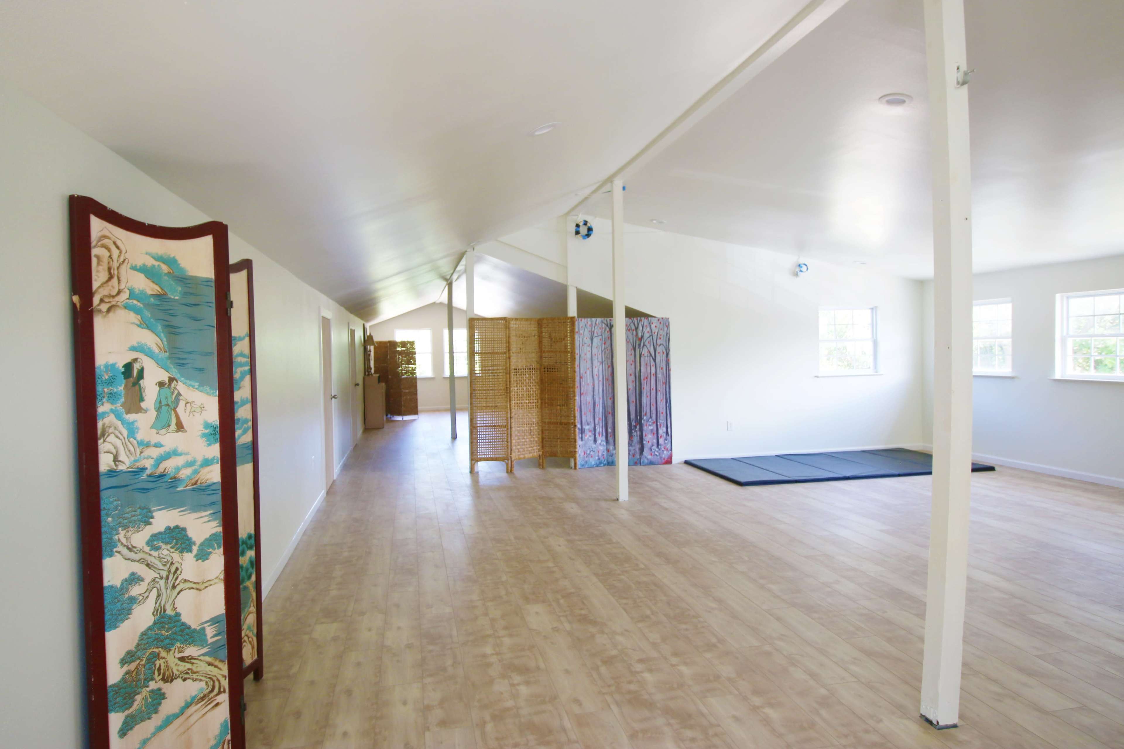 The image shows a spacious, light-filled room designed for yoga or exercise, featuring wooden flooring, folding screens, and mats on the ground.