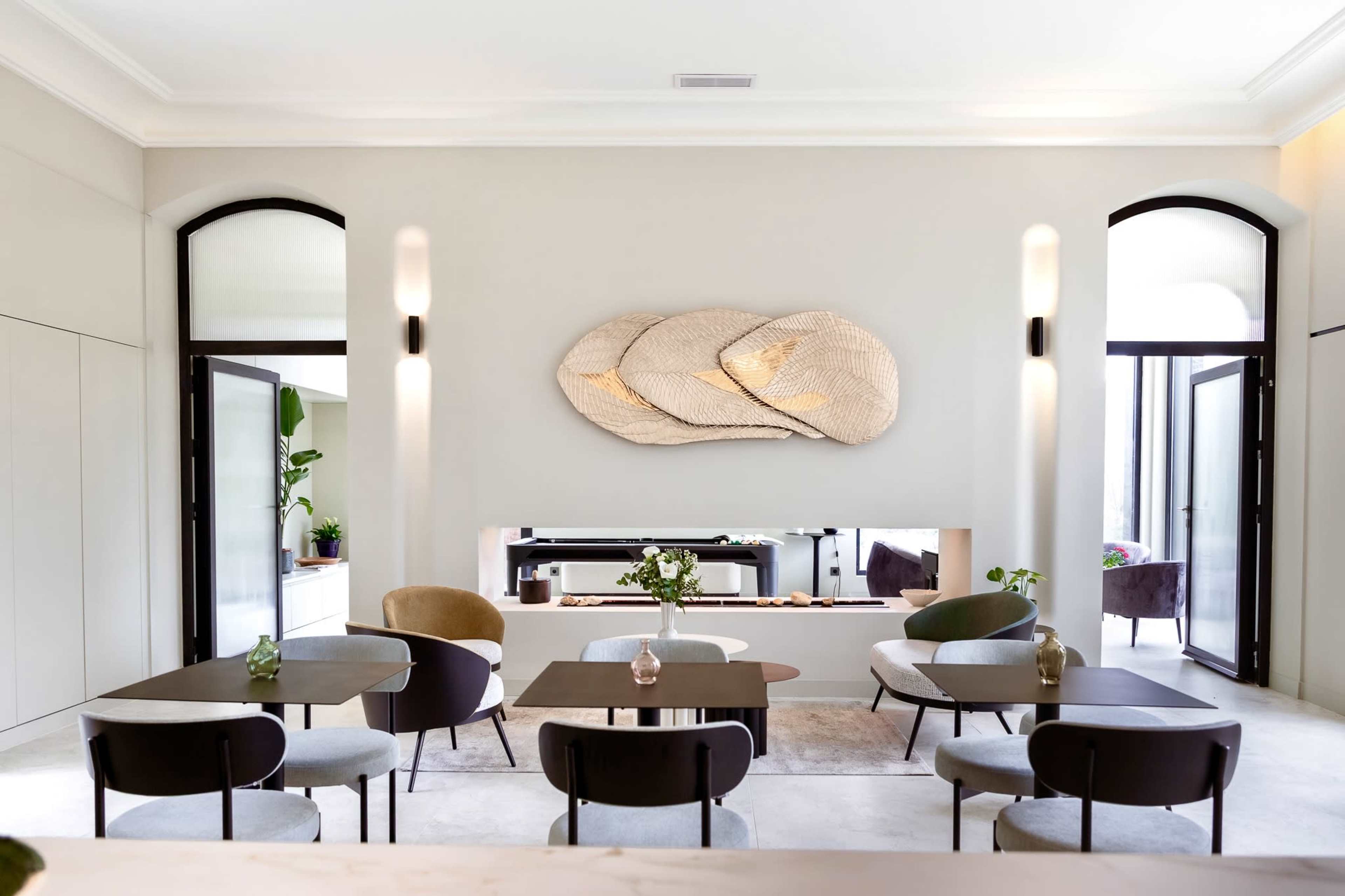 The image shows a modern café interior with several round tables and chairs arranged around a central area, featuring a large decorative wall piece and open doors leading to an outdoor space.