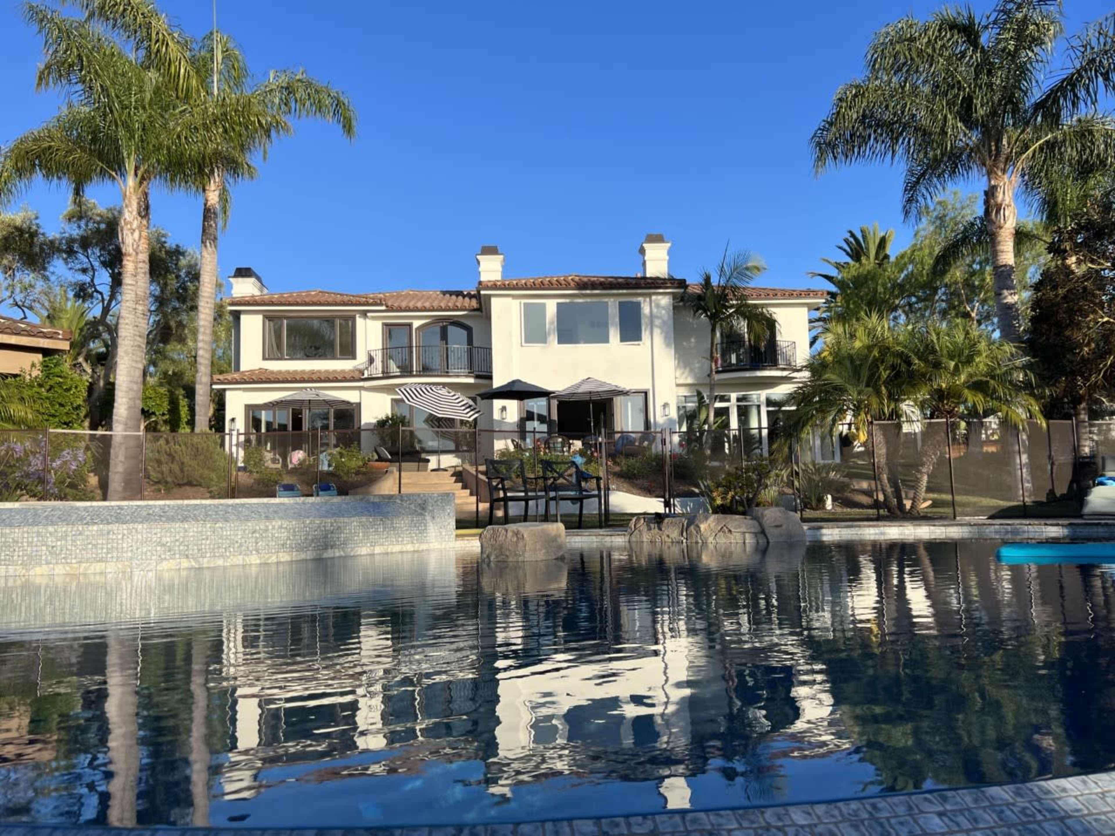 A large two-story house with a pool in the foreground is surrounded by palm trees and landscaped gardens.