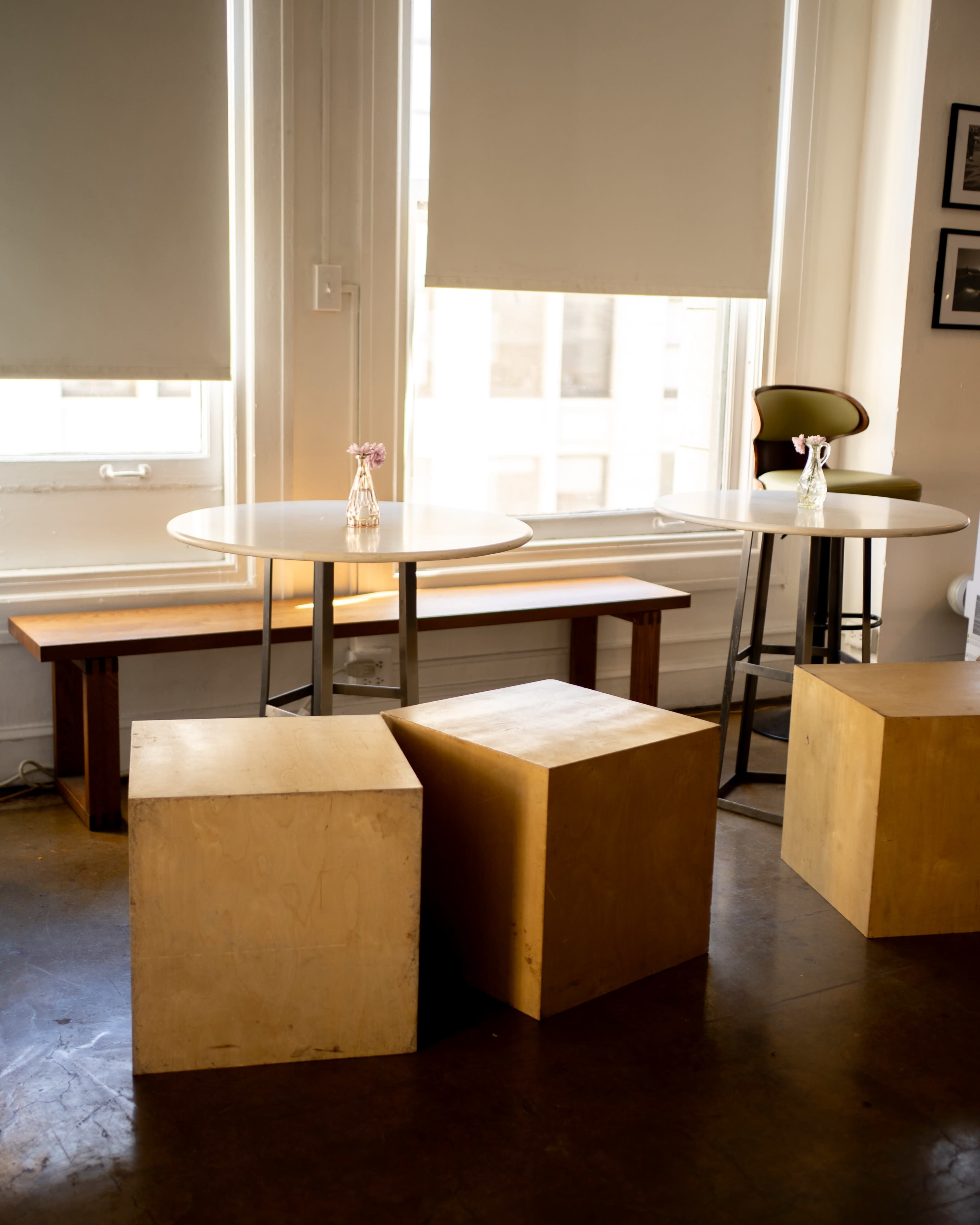 The image shows a small indoor seating area with a round table, a high stool, wooden benches, and cubes, all placed near large windows.