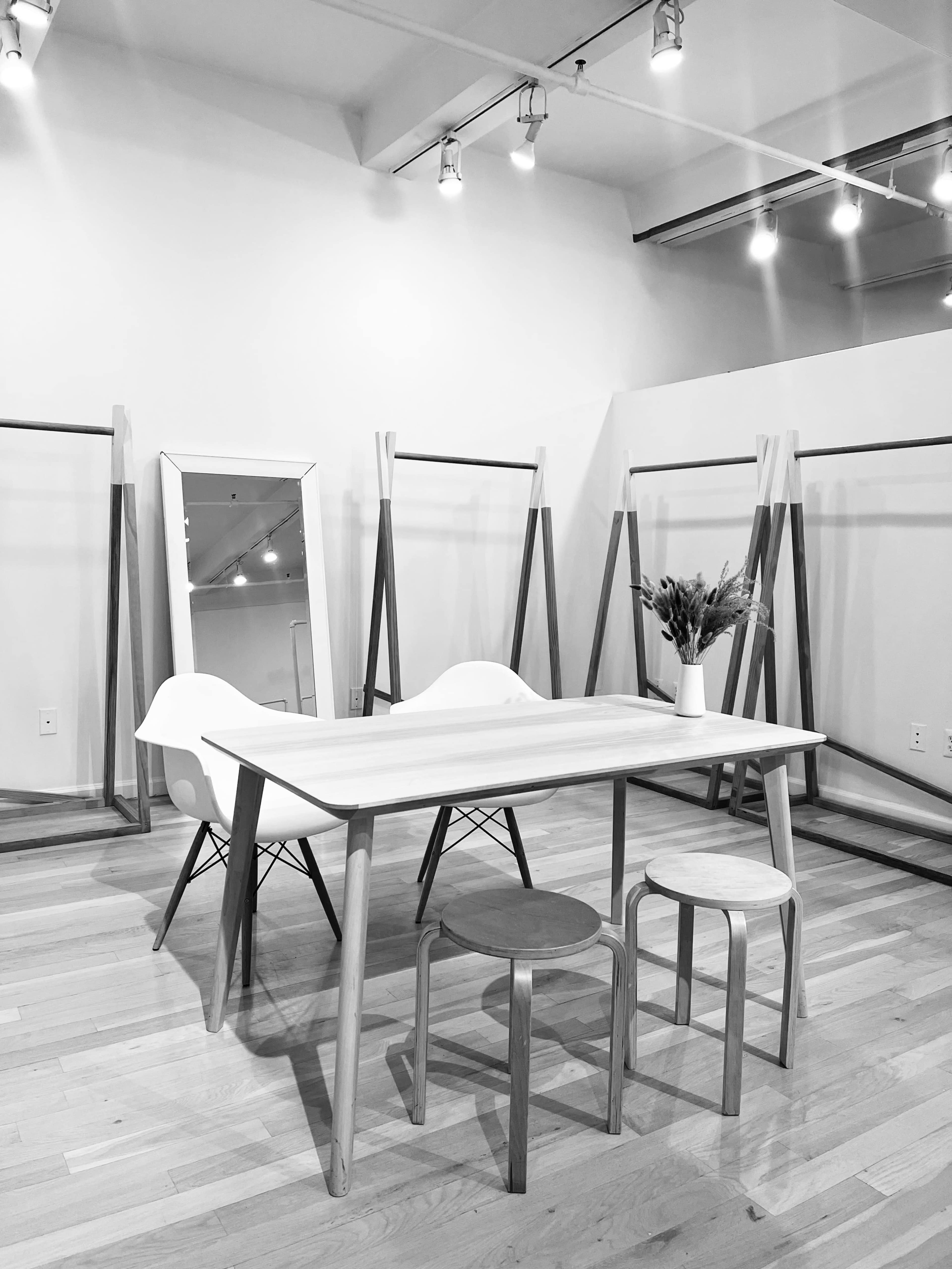 A minimalist room features a wooden table surrounded by two stools and a chair, with mirrors and display racks in the background.