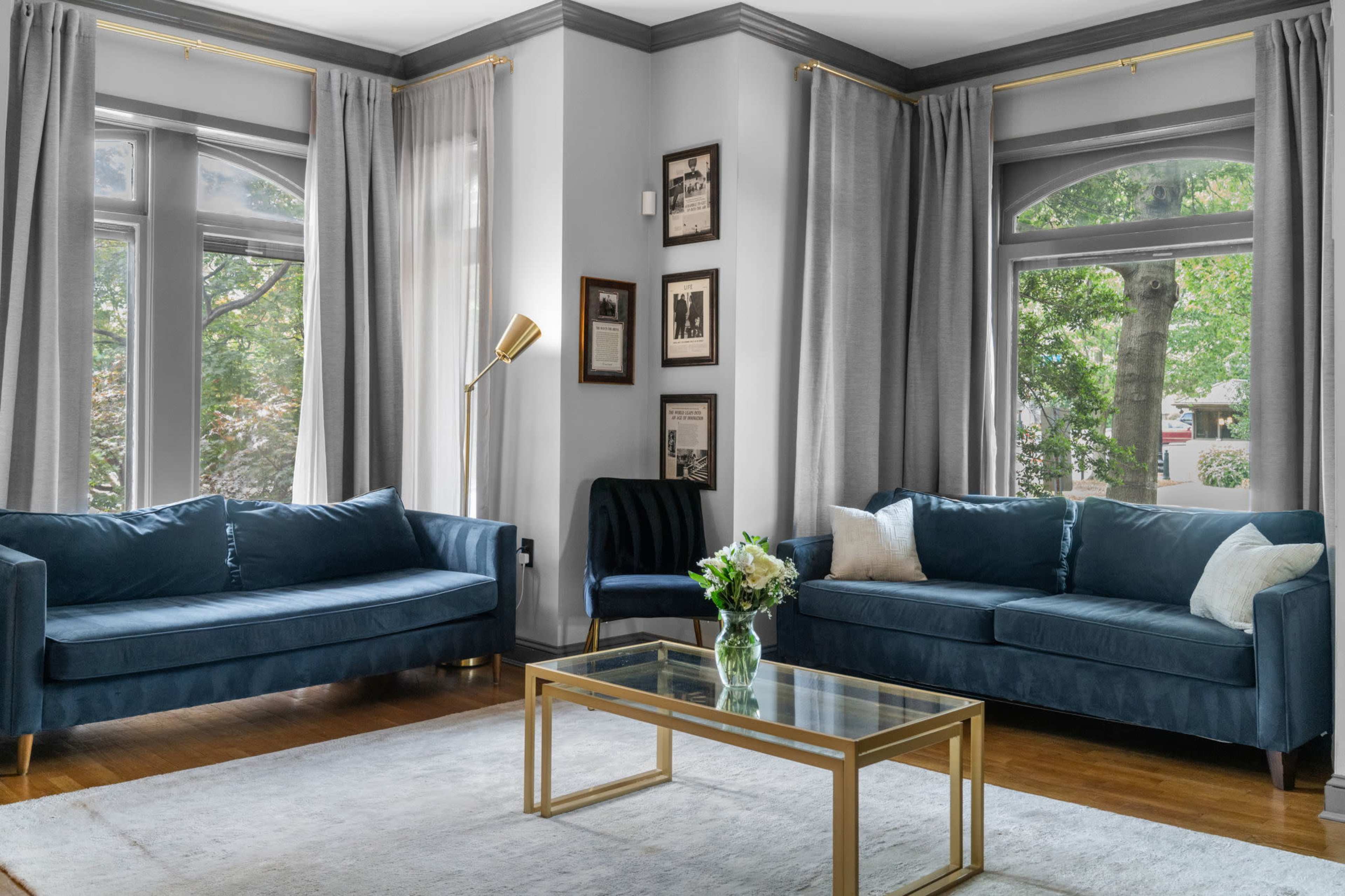 The living room features two blue sofas, a glass coffee table, and large windows with sheer curtains that let in natural light.