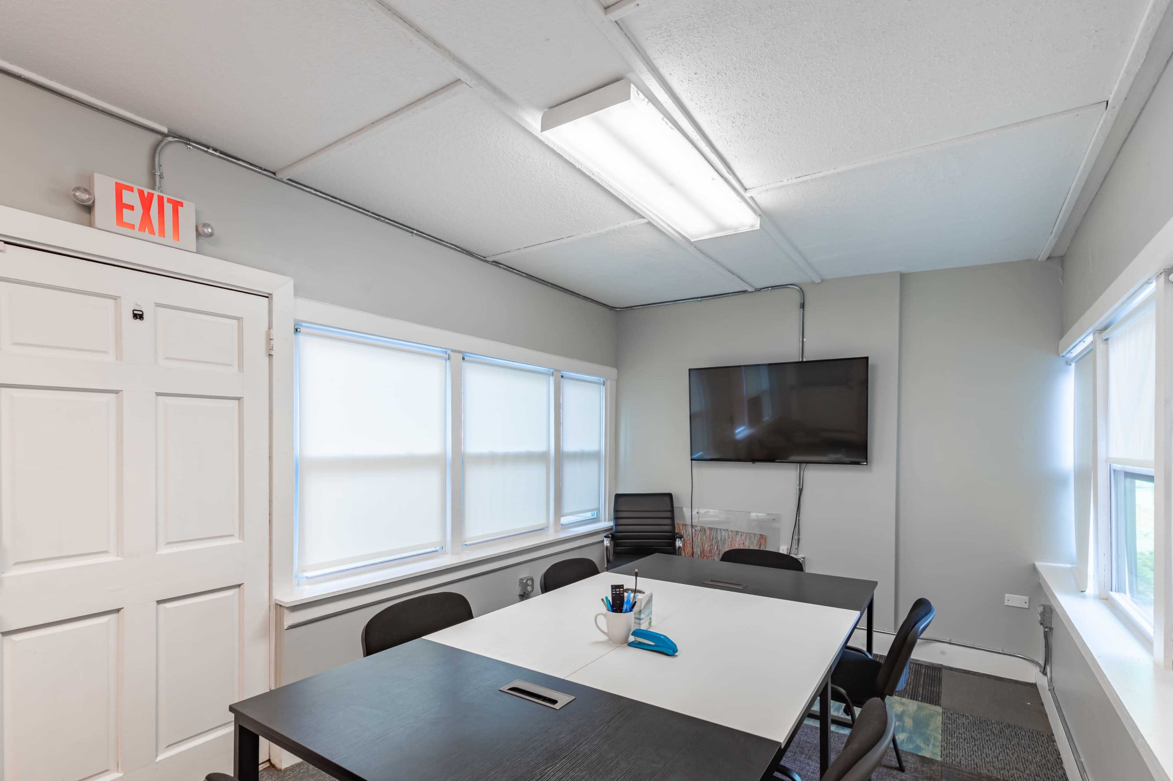 A small conference room features a large table, black chairs, a television mounted on the wall, and exit signage.