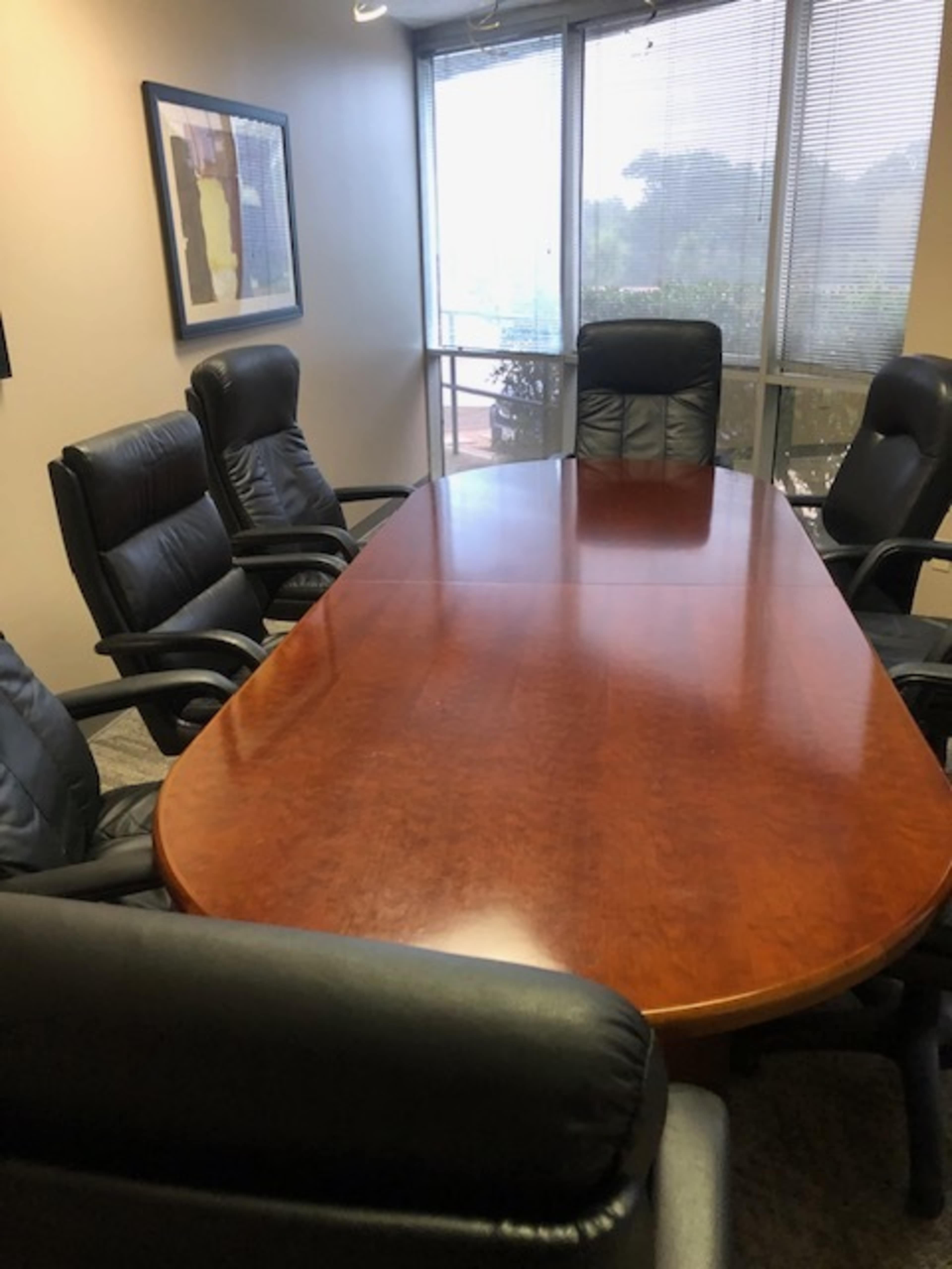 A conference room featuring a large wooden oval table surrounded by six black leather chairs, with windows providing natural light.