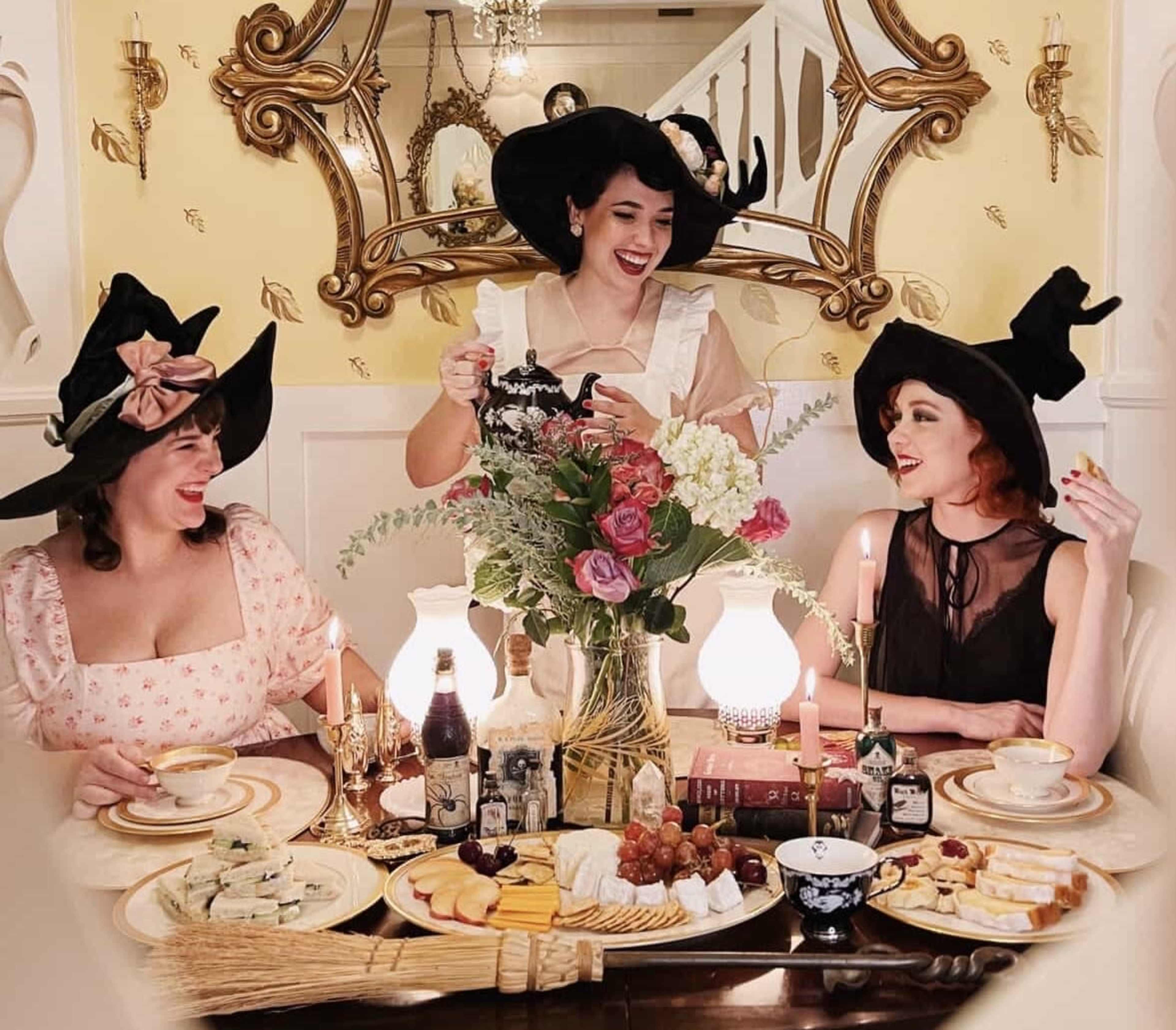 Three women in large black hats gather around a festive table adorned with a variety of food, flowers, and candles, while one serves tea.
