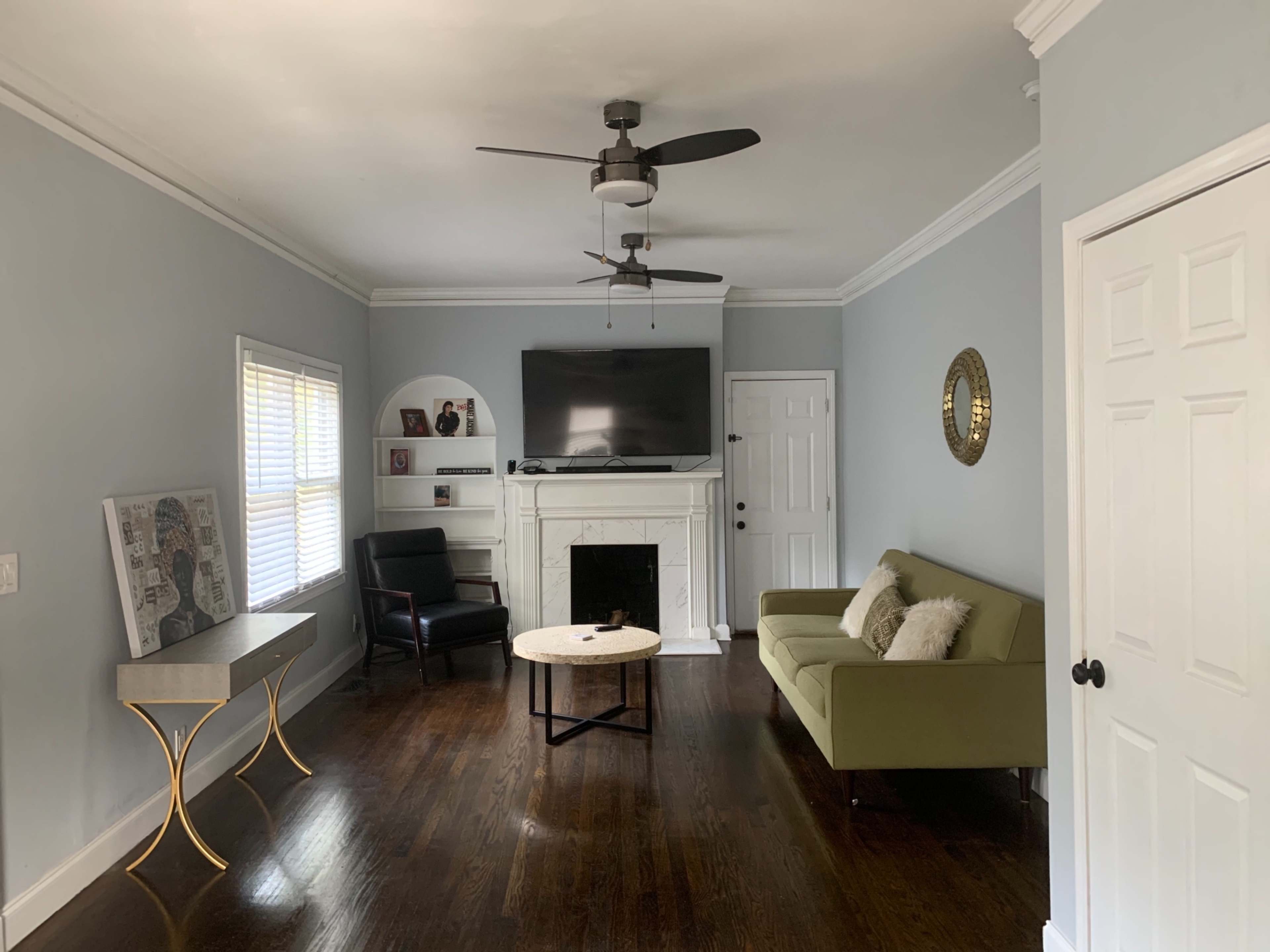 The room features a green sofa, a black chair, a coffee table, and a mounted television above a white fireplace, all set against light gray walls with wooden flooring.