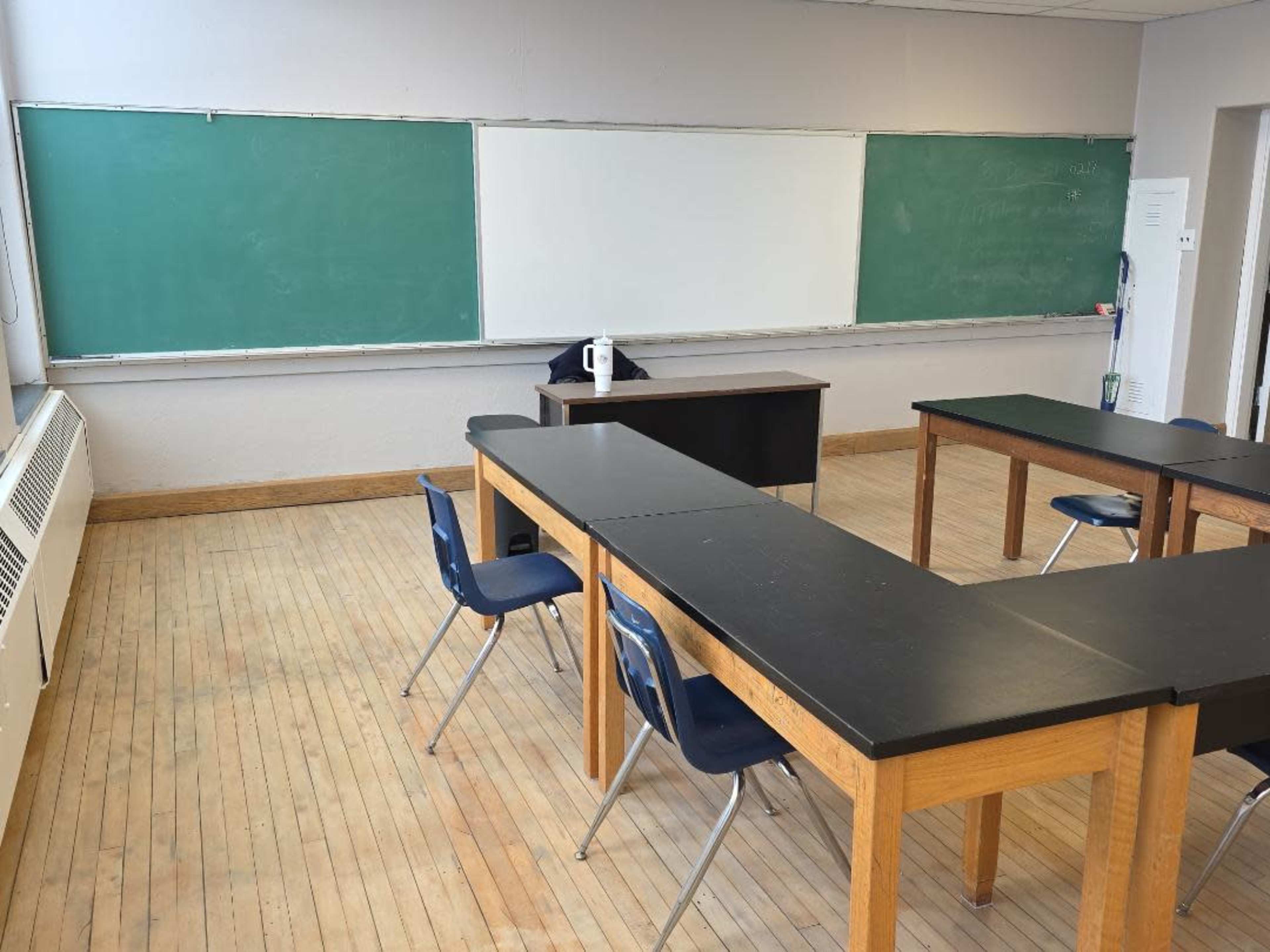 The image shows a classroom with two green chalkboards, a whiteboard, and several wooden tables arranged with blue chairs.