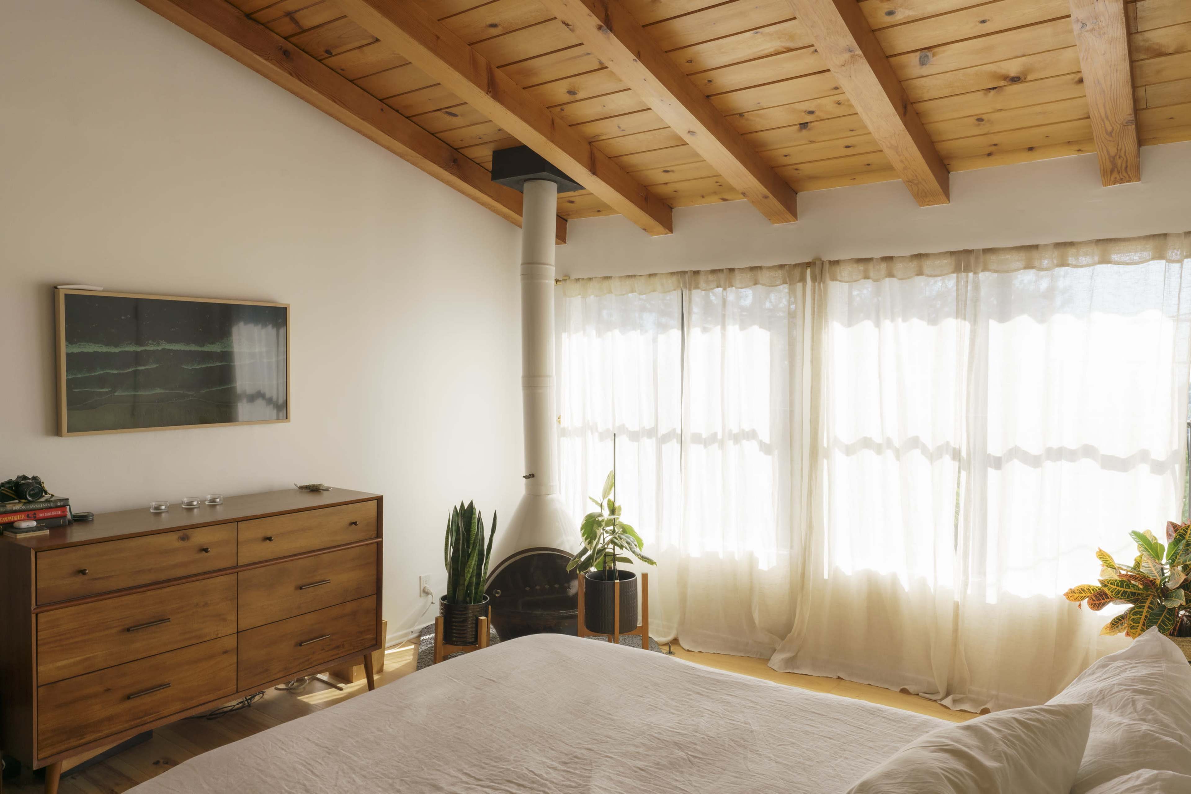A bright bedroom features a wooden ceiling, a dresser, and large windows covered with sheer curtains that allow natural light to enter.