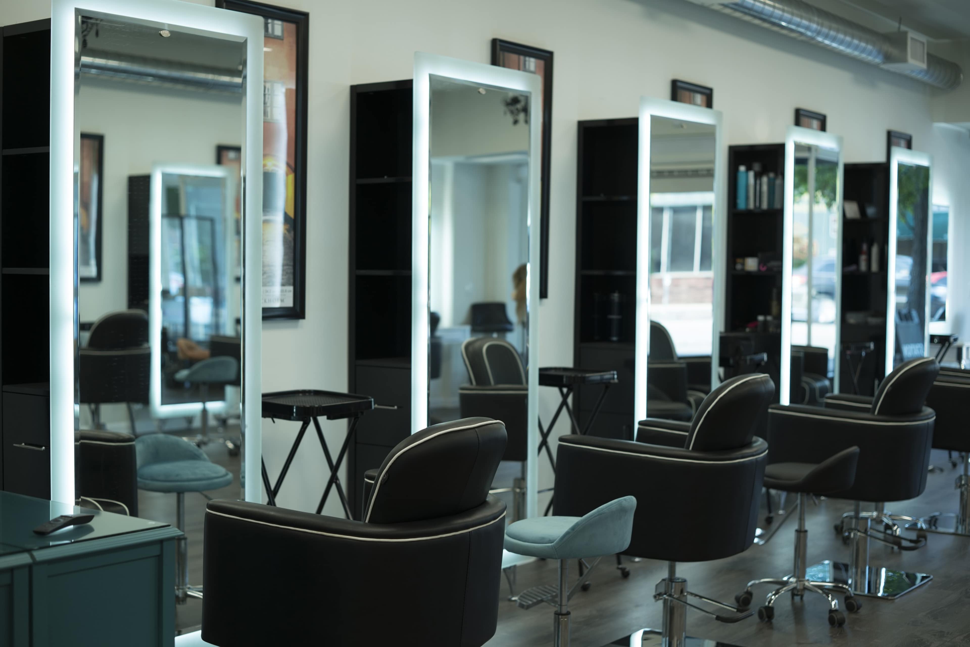 A hair salon with several styling stations, each featuring illuminated mirrors and black chairs arranged in a row.