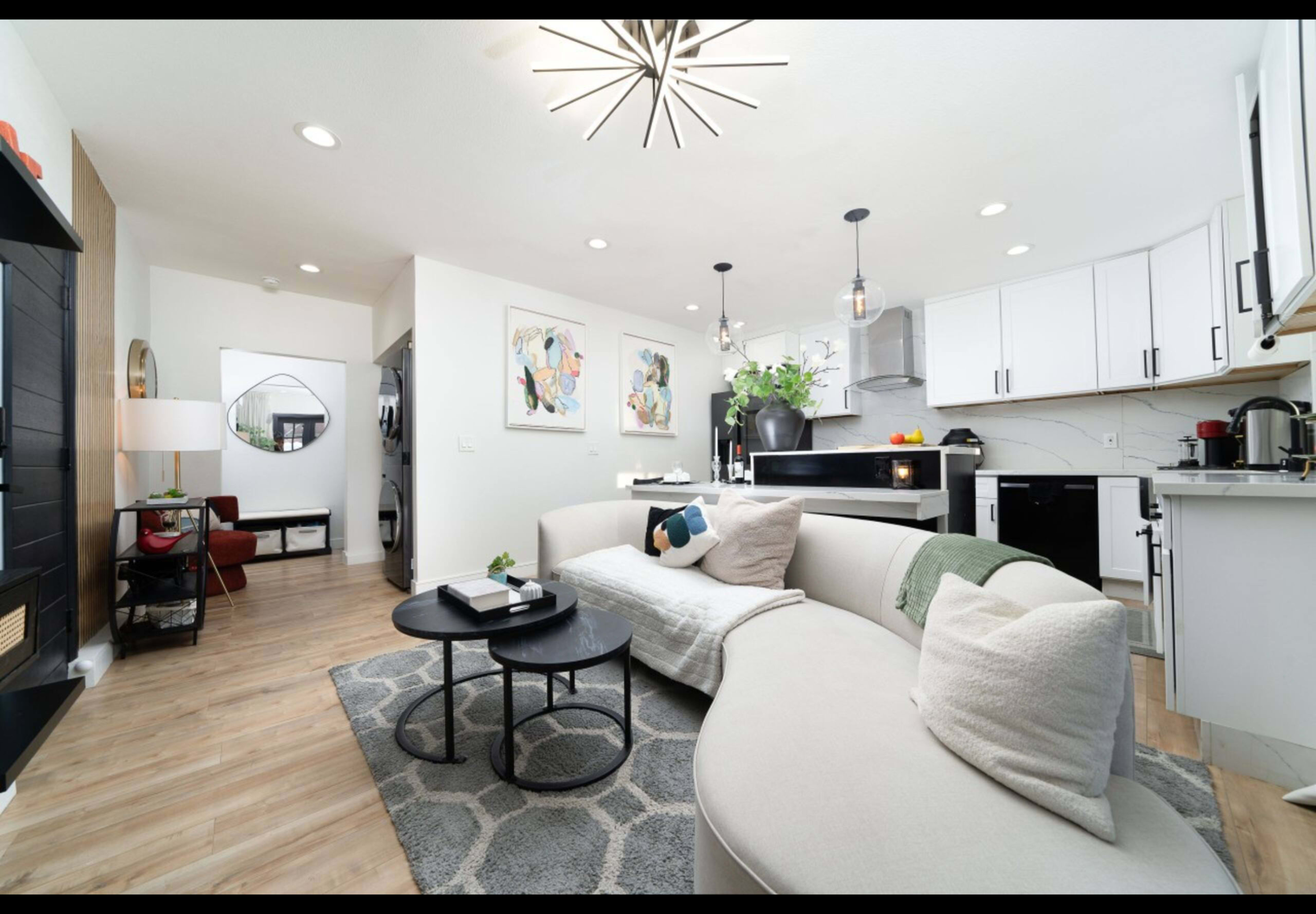 A modern living area featuring a curved sectional sofa, a coffee table, and a well-equipped kitchen with white cabinetry and pendant lighting.