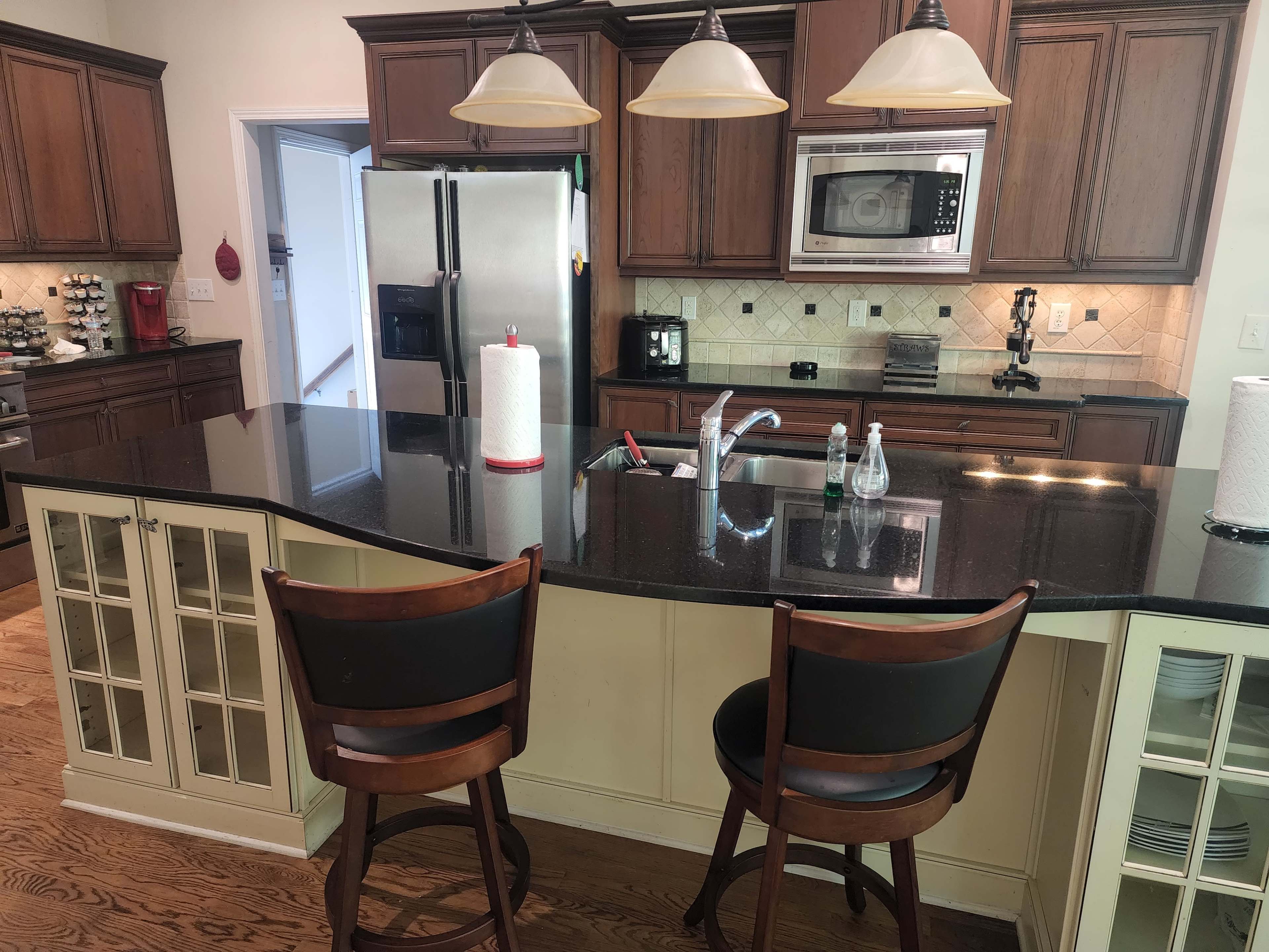 A modern kitchen features dark wooden cabinets, a granite countertop island with two bar stools, and stainless steel appliances.