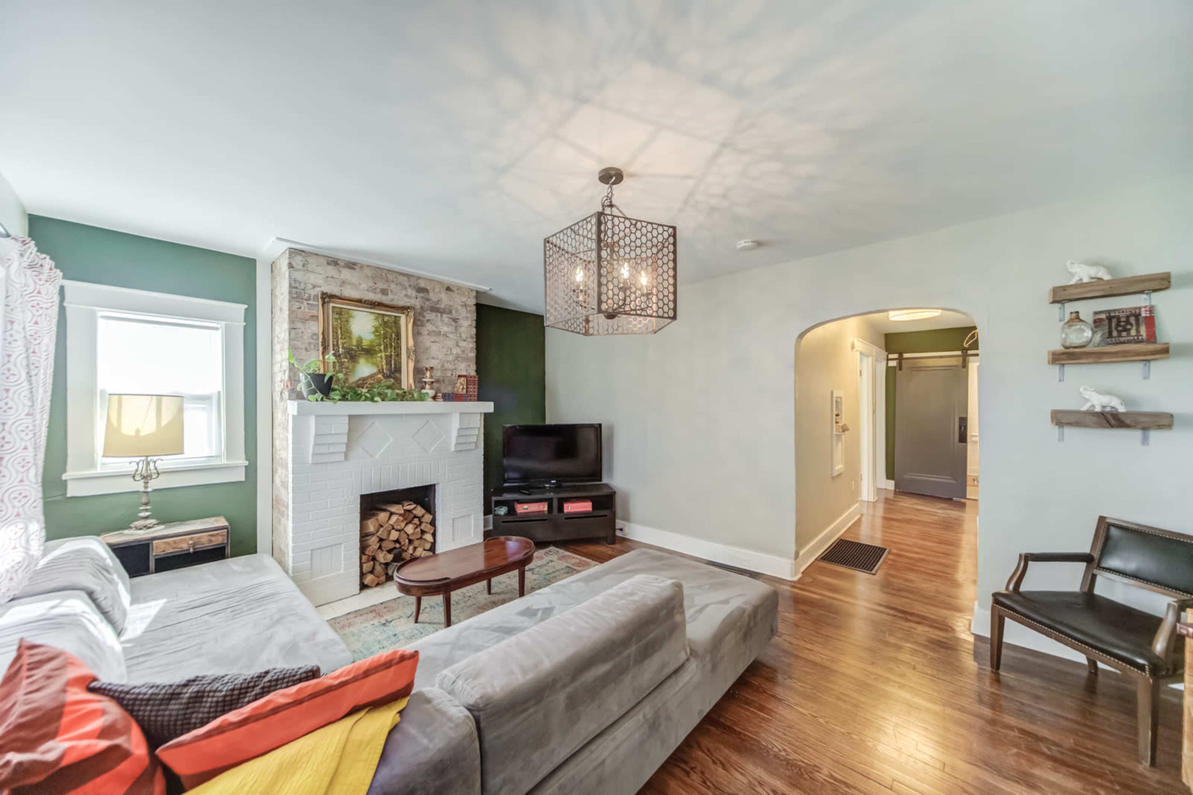 The living room features a gray sectional sofa, a fireplace with stacked firewood, and a television against a green accent wall.
