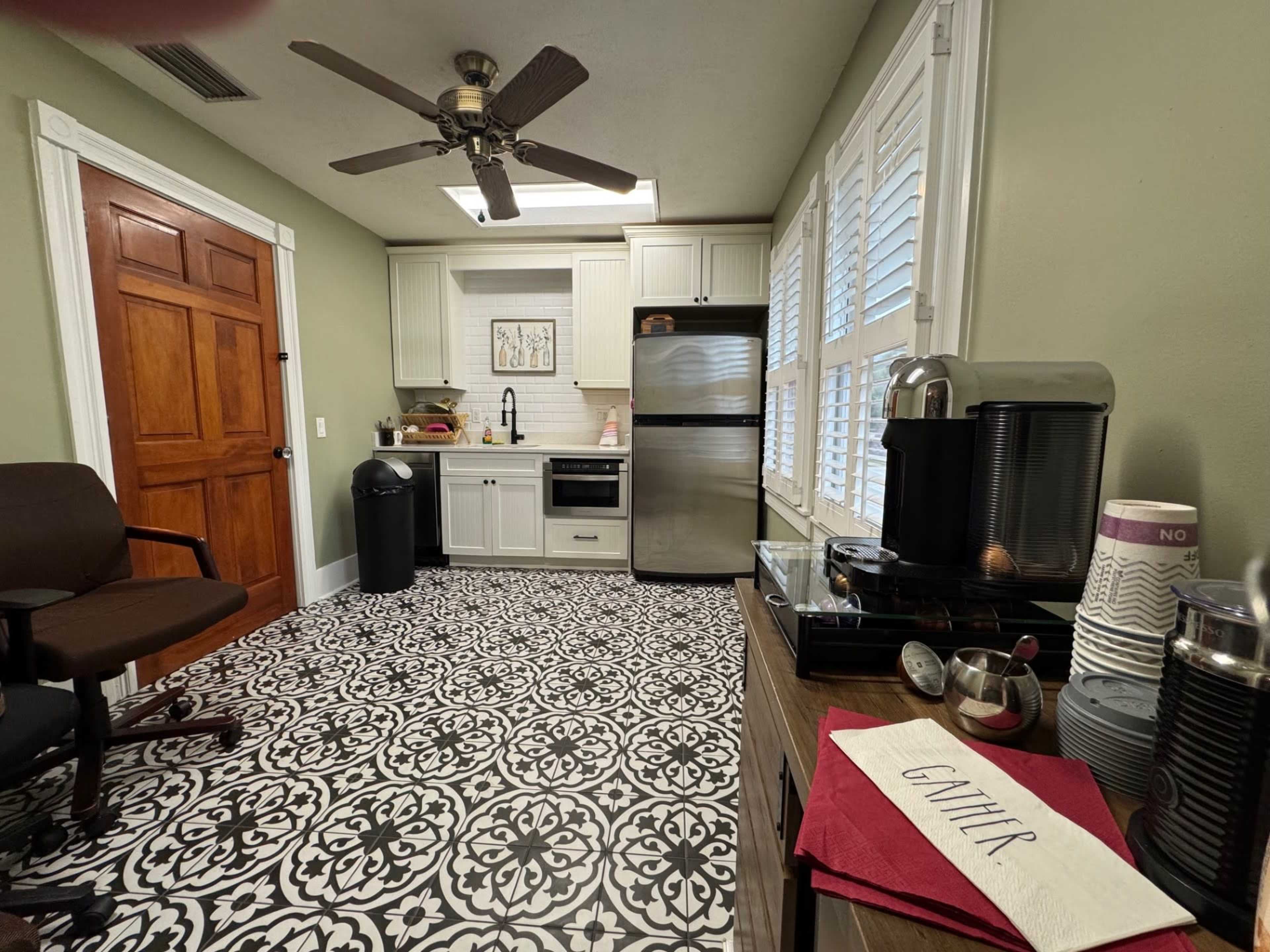 The image shows a kitchen with a black and white patterned tile floor, stainless steel appliances, and a coffee station, featuring a fan overhead and wooden elements in the cabinetry and door.