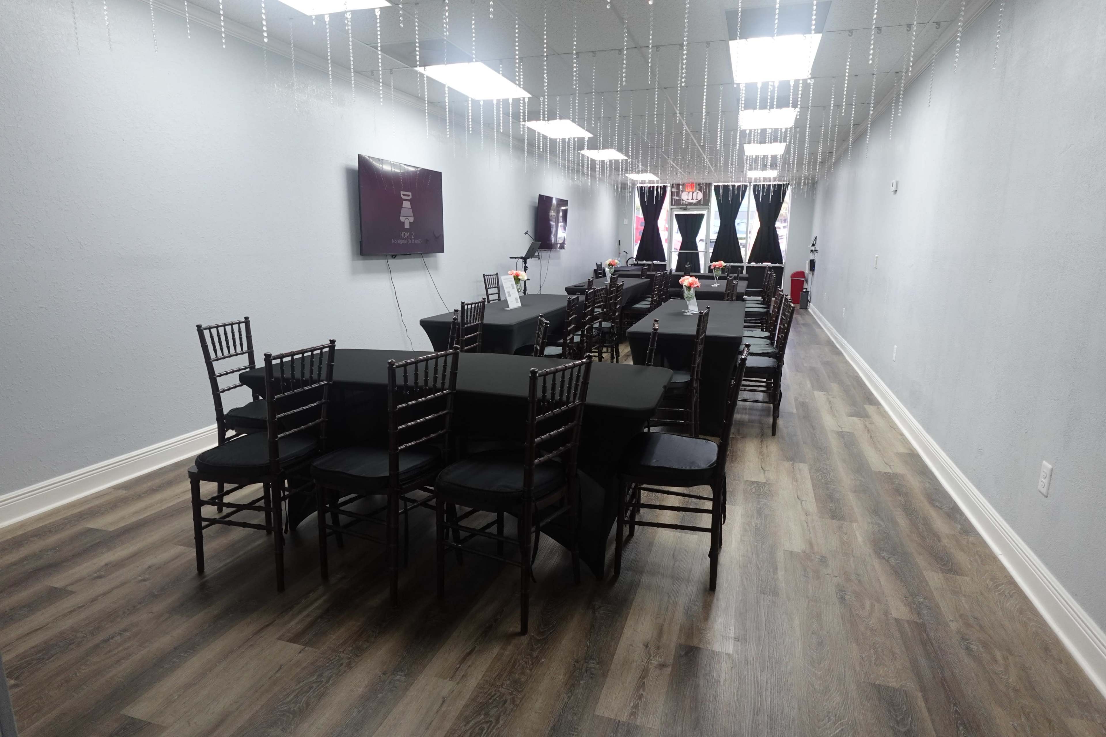 A spacious room neatly arranged with black-covered tables and chairs, featuring a light-colored wooden floor and decorative lighting overhead.