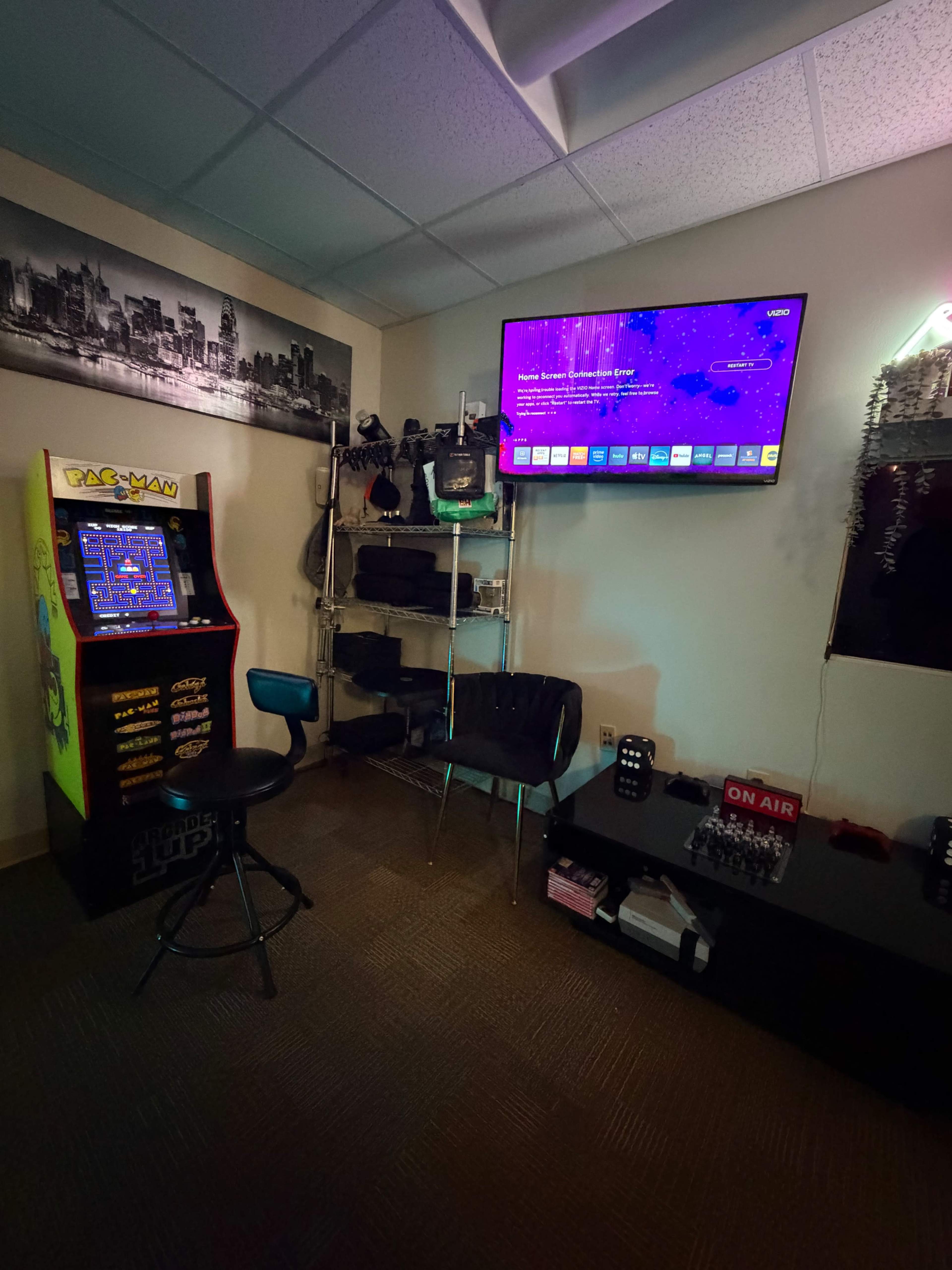 The image shows a gaming room featuring a Pac-Man arcade machine, a television displaying a colorful interface, and various shelves with memorabilia and equipment.