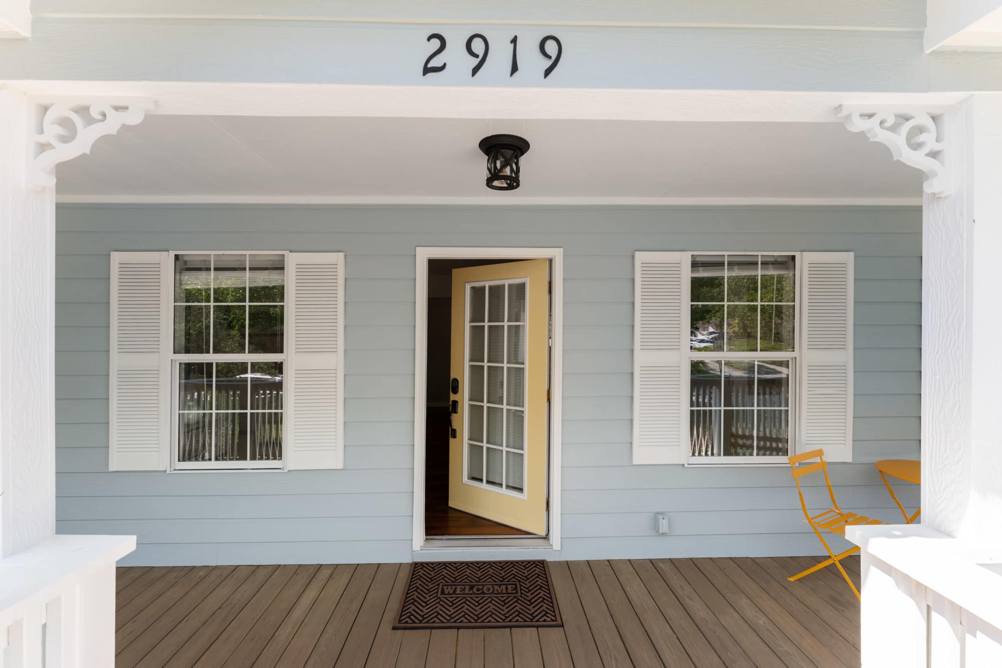 A front porch of a light blue house with a yellow door, two white windows, and an orange chair, featuring a welcome mat at the entrance.