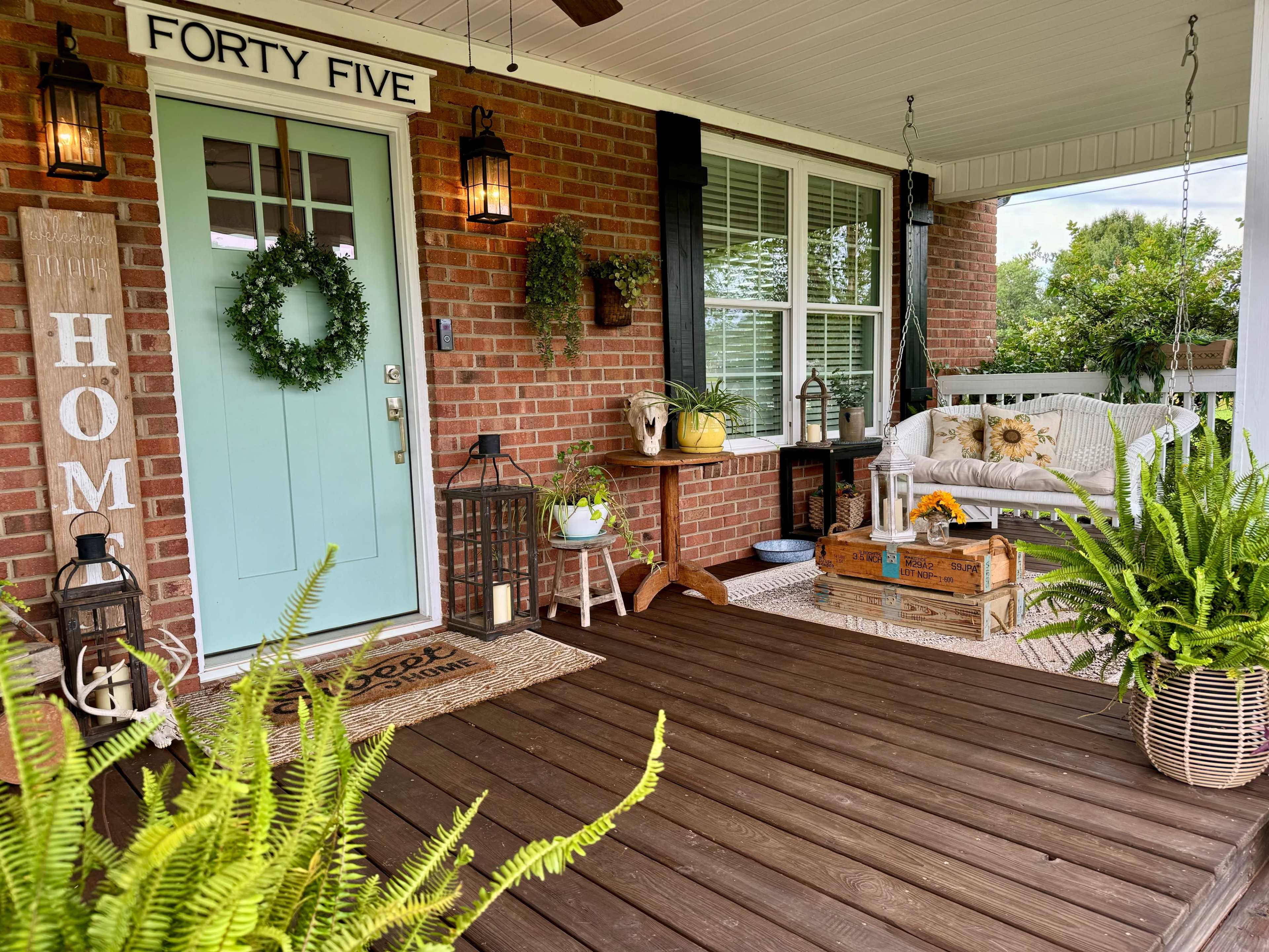 A welcoming porch area with a light blue door, a decorative wreath, and various plants arranged around a swing and wooden furniture on a wooden deck.