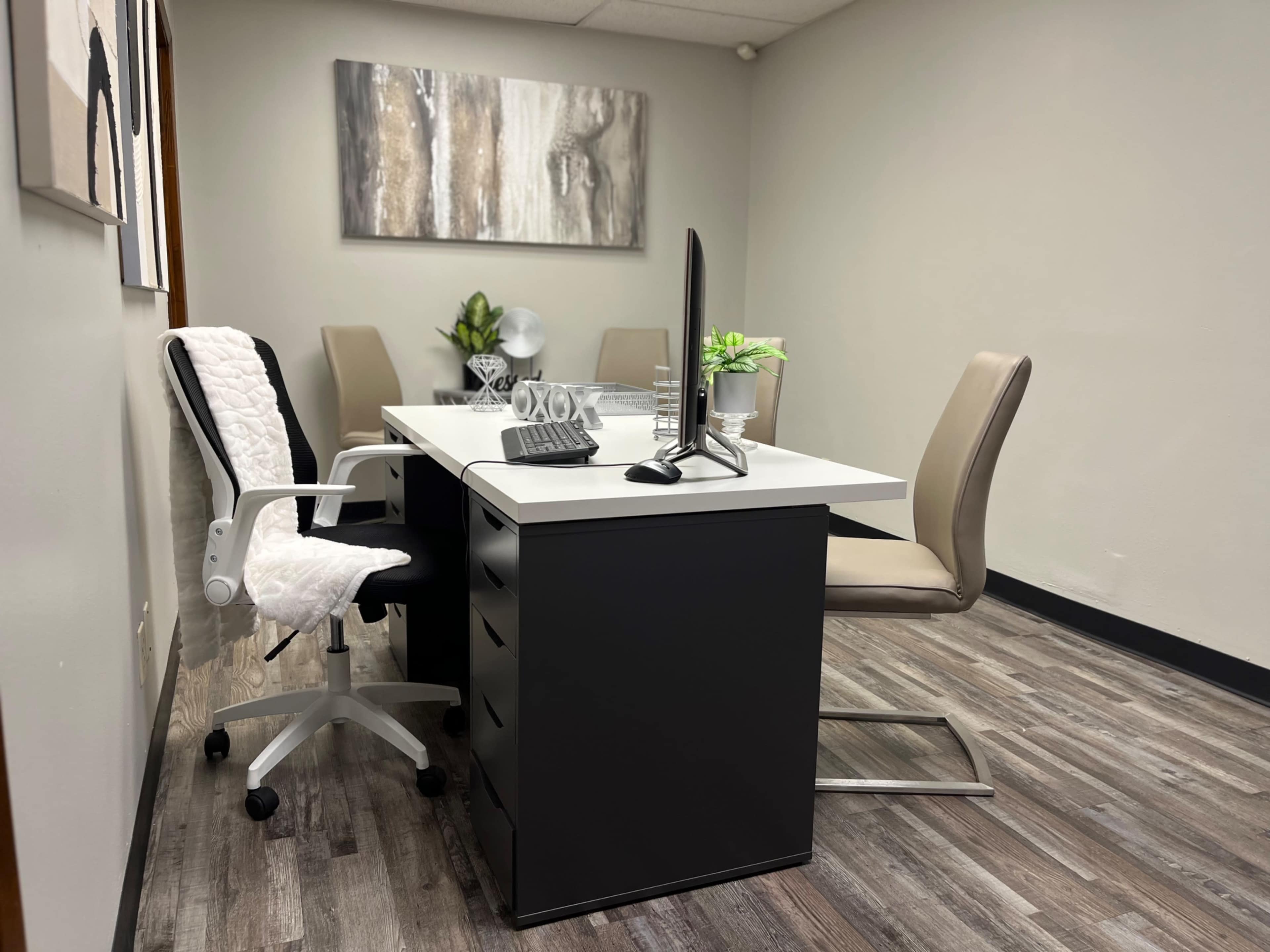 A modern office space featuring a white desk with a computer monitor, surrounded by two chairs and decorated with small plants.