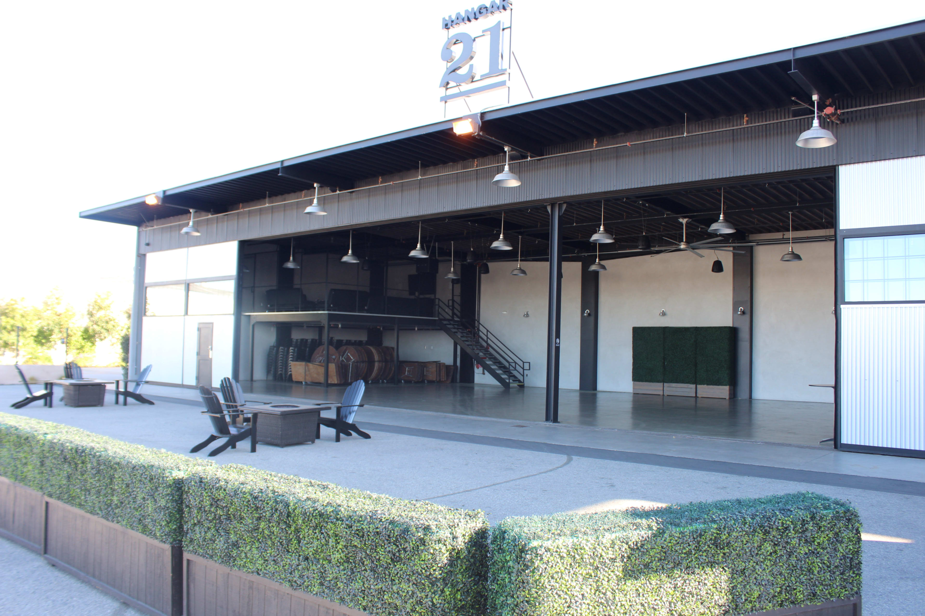 A large, open building with a tall ceiling, featuring a staircase and rows of barrels in the background, surrounded by a neatly trimmed hedge in the foreground.