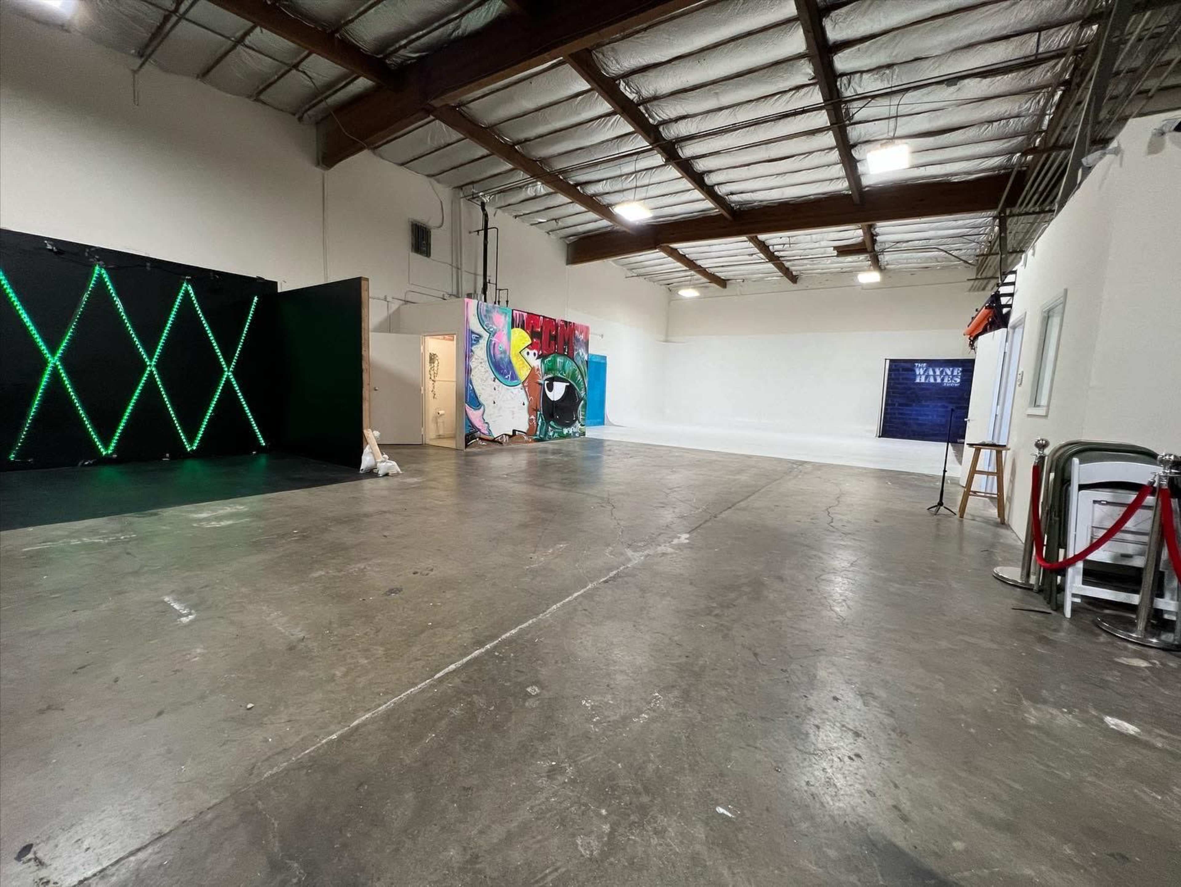 A spacious indoor area with concrete flooring, featuring a wall with neon lights, colorful graffiti art, and a simple backdrop along with a few props.