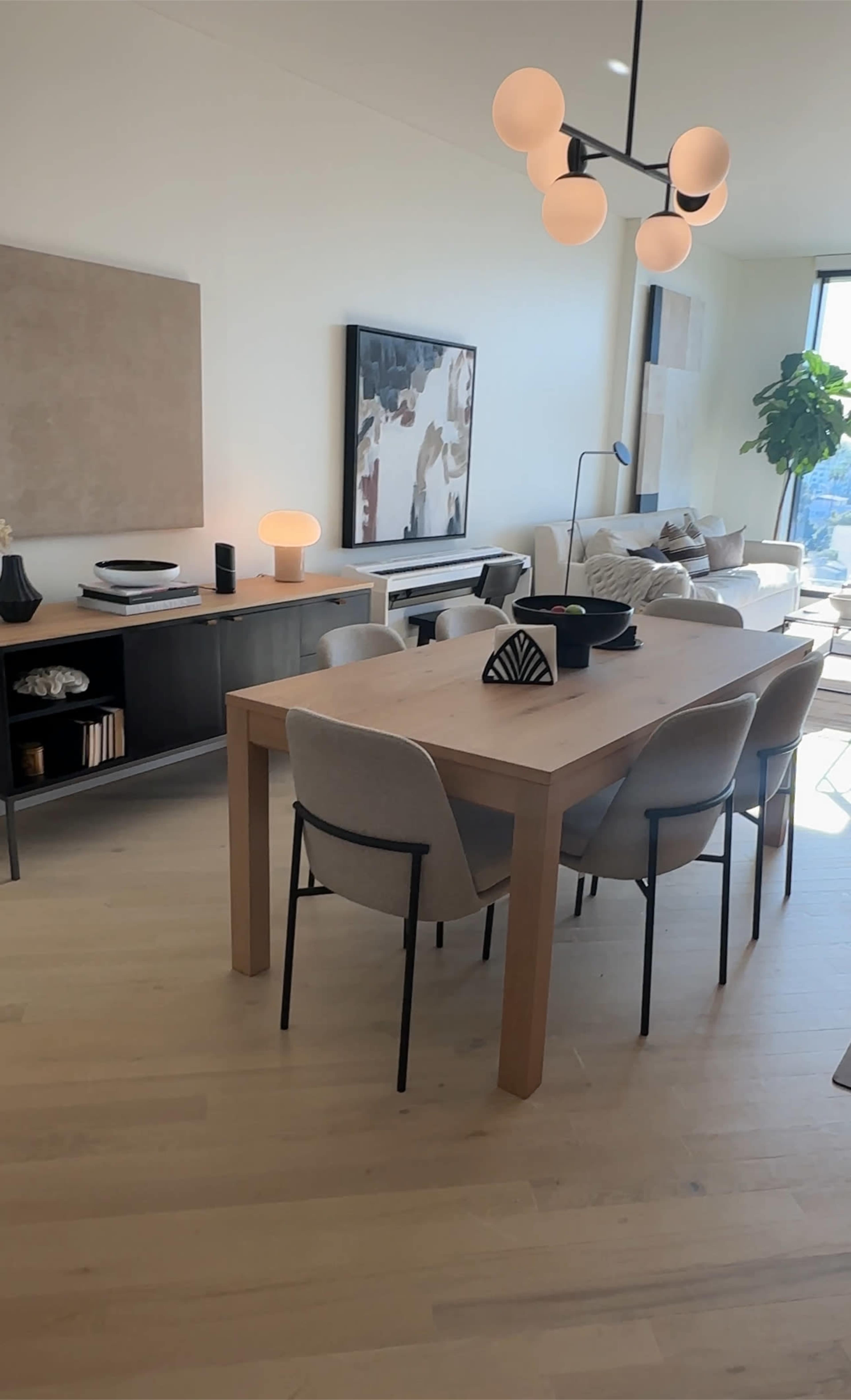 A modern dining area features a wooden table surrounded by upholstered chairs, with a sideboard and large windows allowing natural light to fill the space.