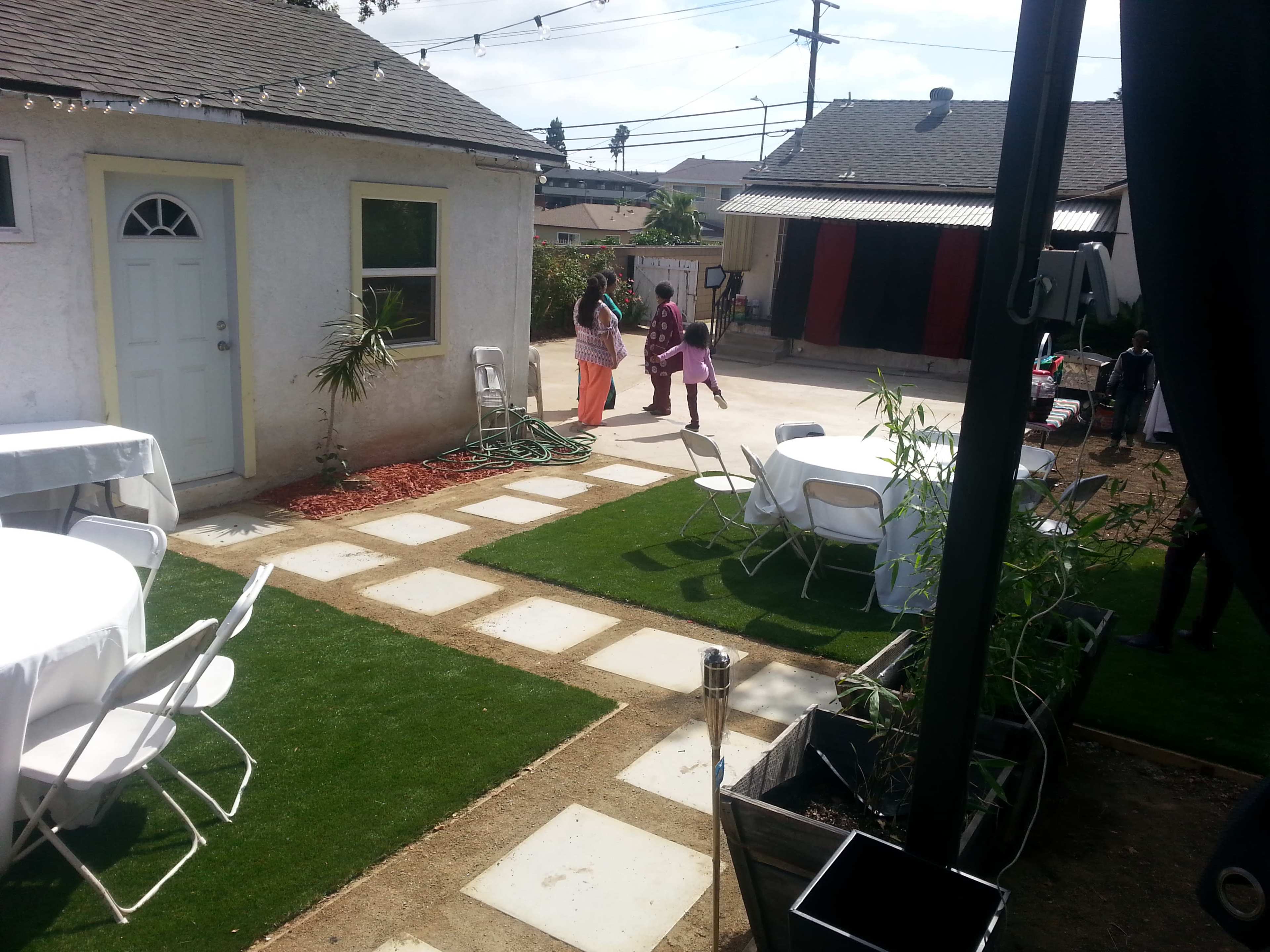 A backyard area features neatly arranged tables and chairs, with a small group of people walking near a house and an adjacent building.
