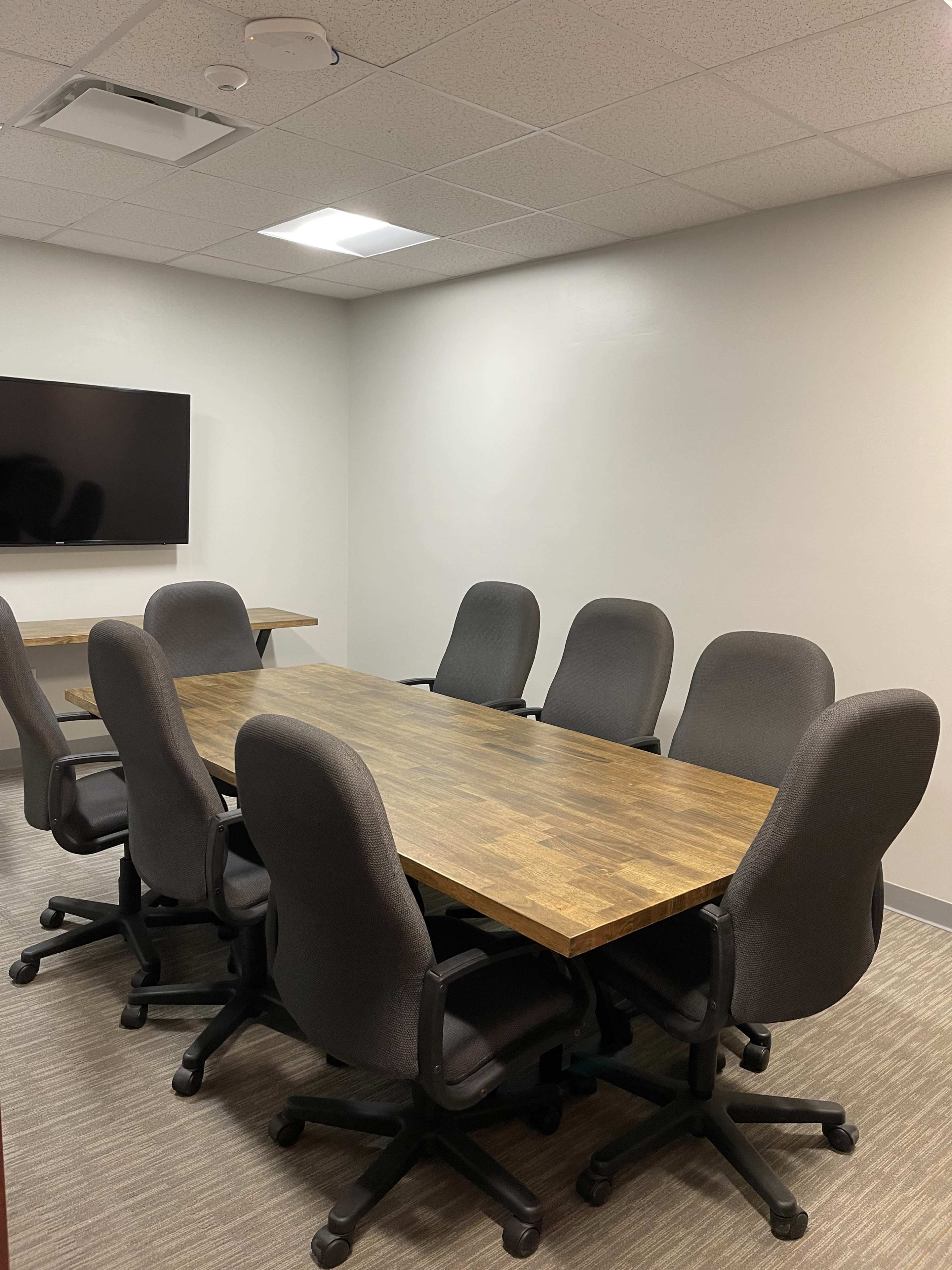 A conference room features a large wooden table surrounded by six ergonomic chairs, with a wall-mounted screen in the background.