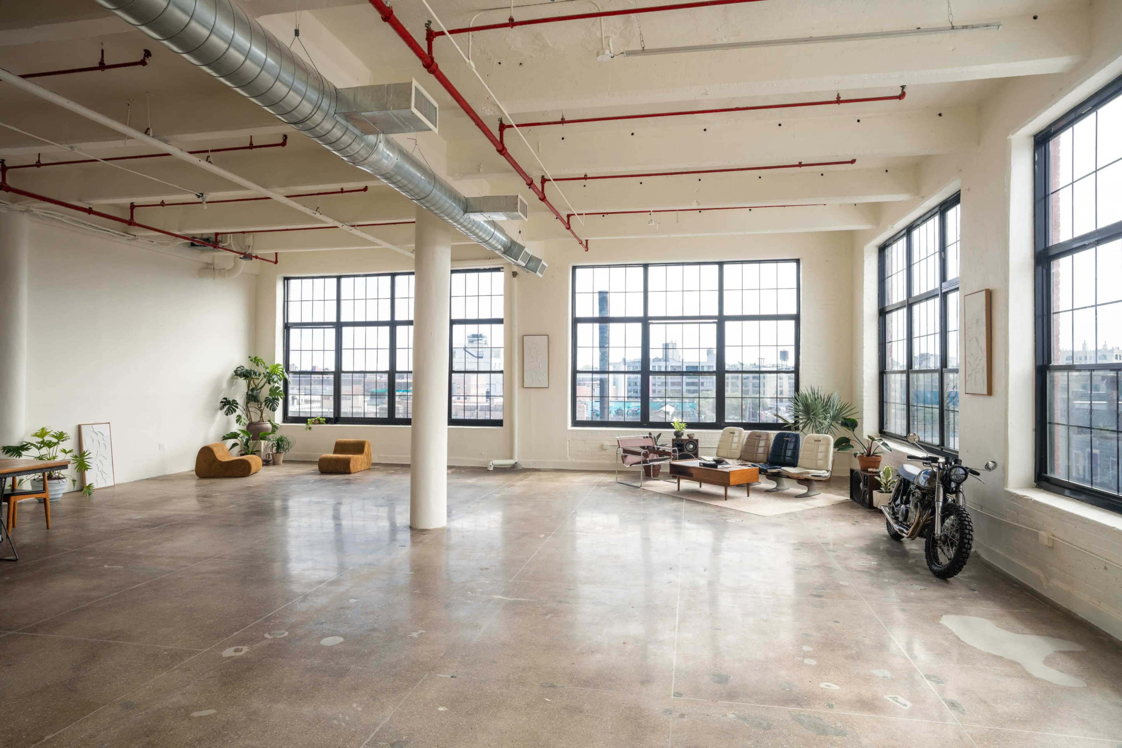 The image shows an expansive, empty industrial-style room with large windows, concrete flooring, and minimal furnishings, including plants and seating arrangements.