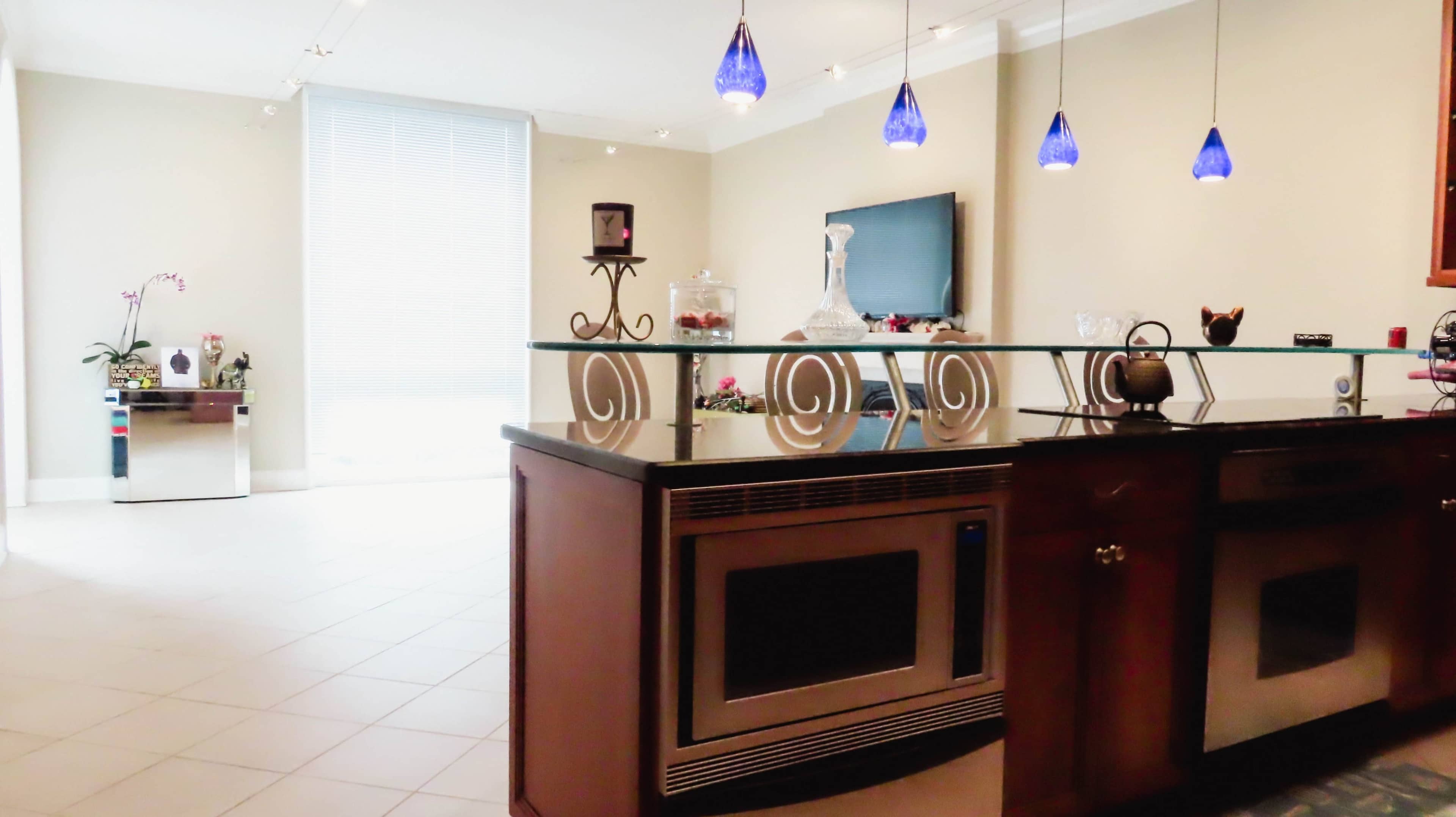 A modern kitchen area with a glass partition, dark countertops, and blue pendant lights hanging above.