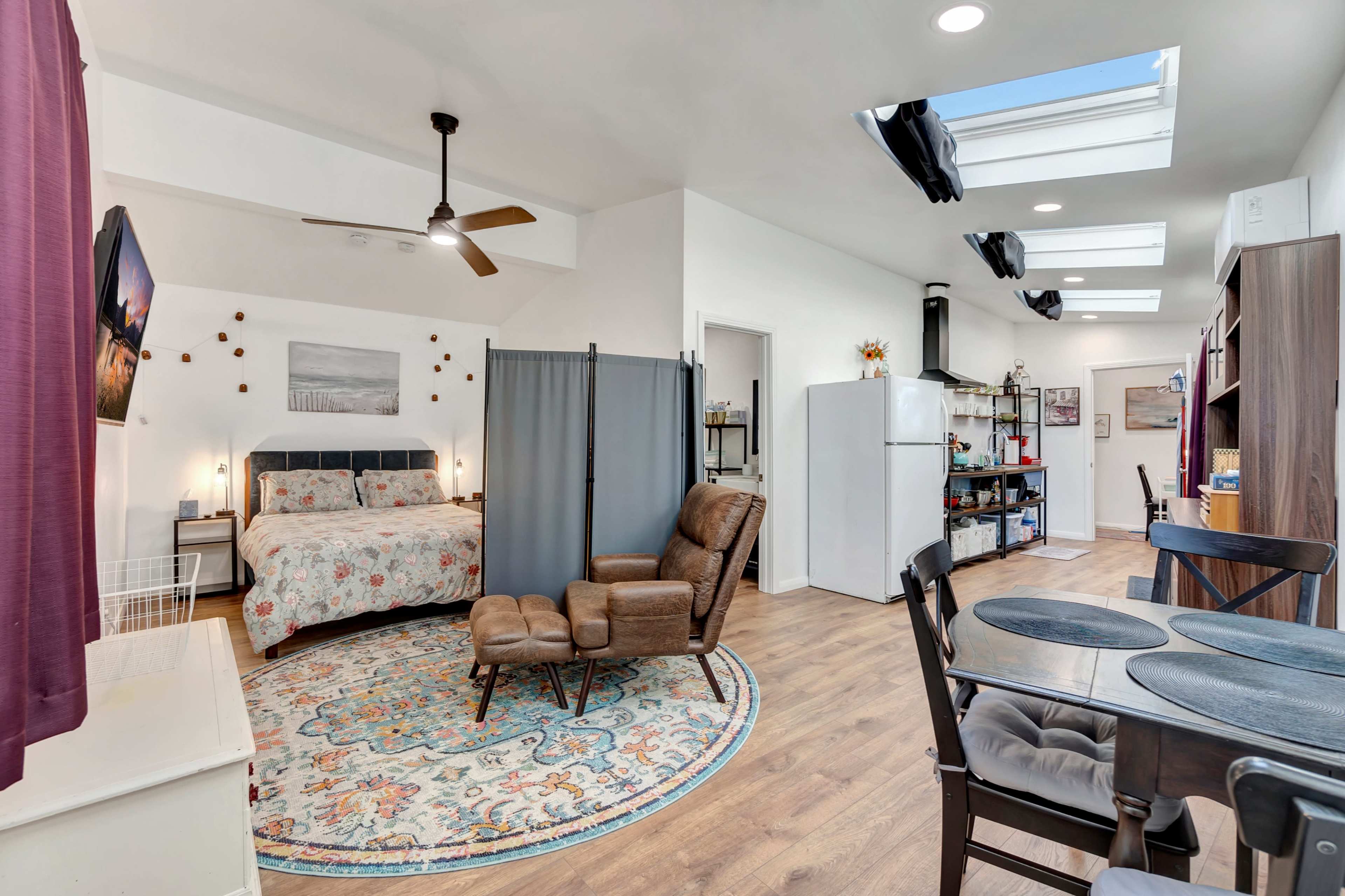 The image shows a small, open-concept living space featuring a bed, seating area with a recliner, a dining table, and a kitchenette.