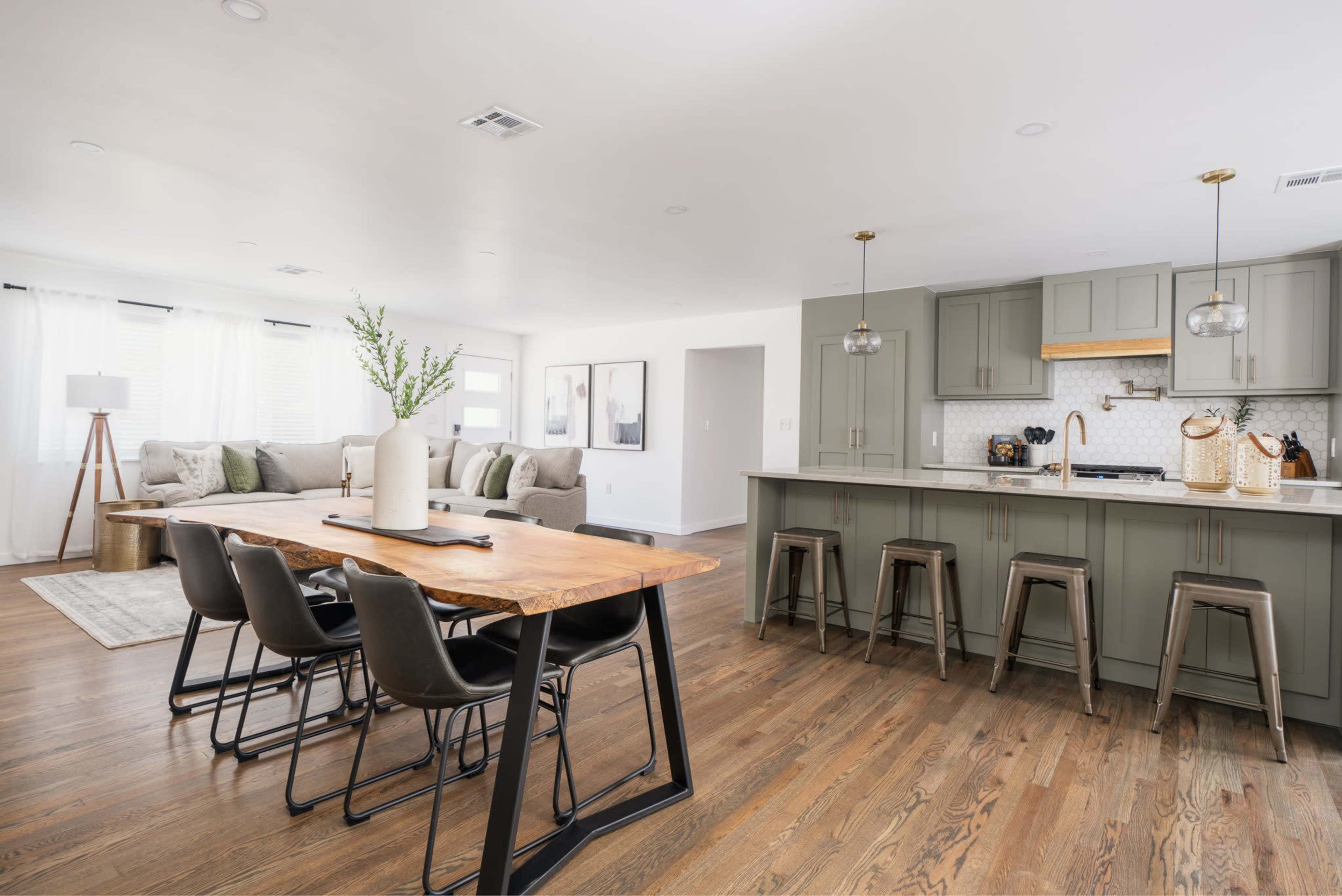 A modern open-concept living space featuring a dining area with a wooden table and black chairs adjacent to a kitchen with green cabinets and a white backsplash.