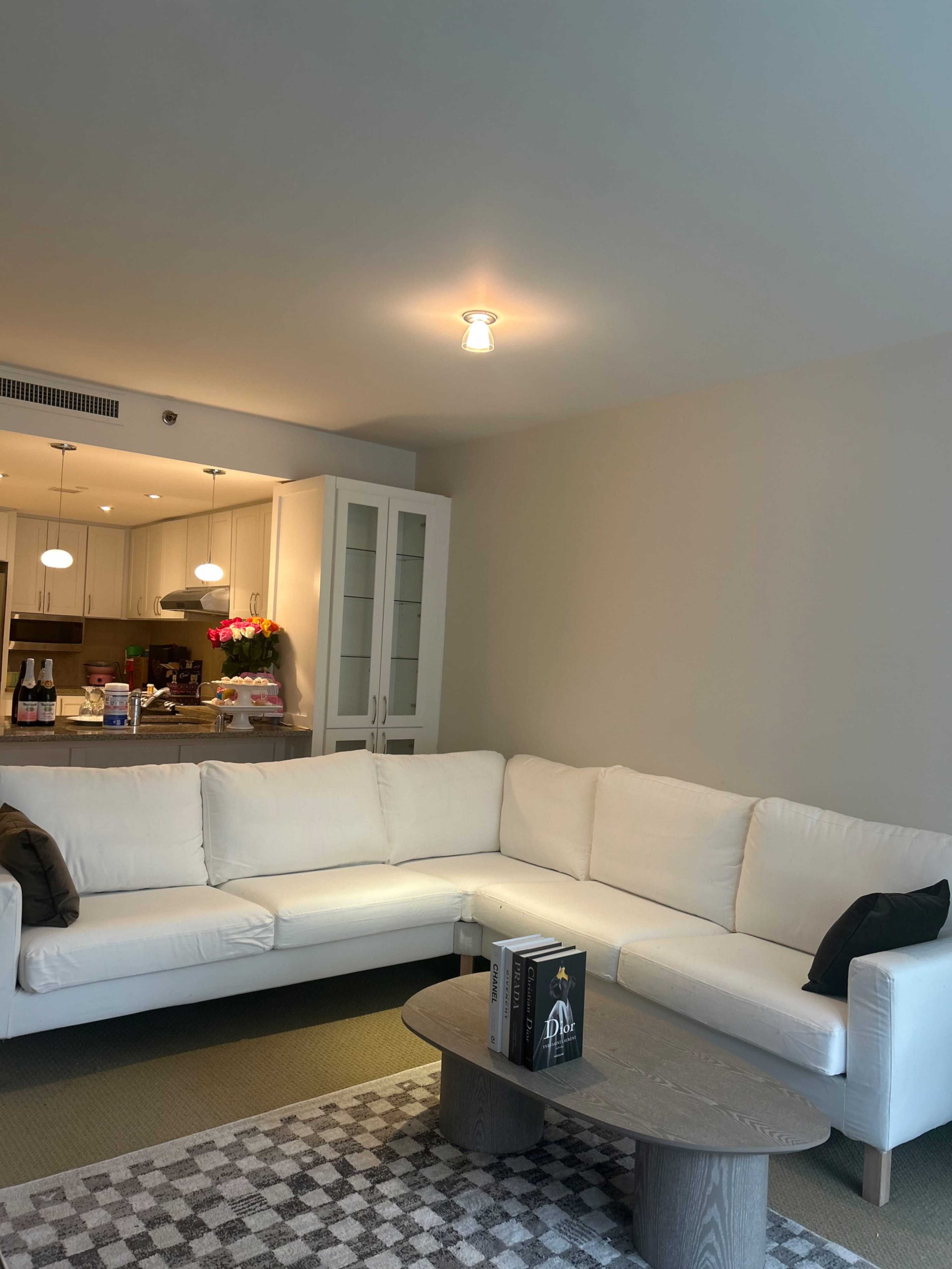 A light-colored, modern sectional sofa is positioned in a living room with a rug, a round coffee table, and a view of a kitchen in the background.