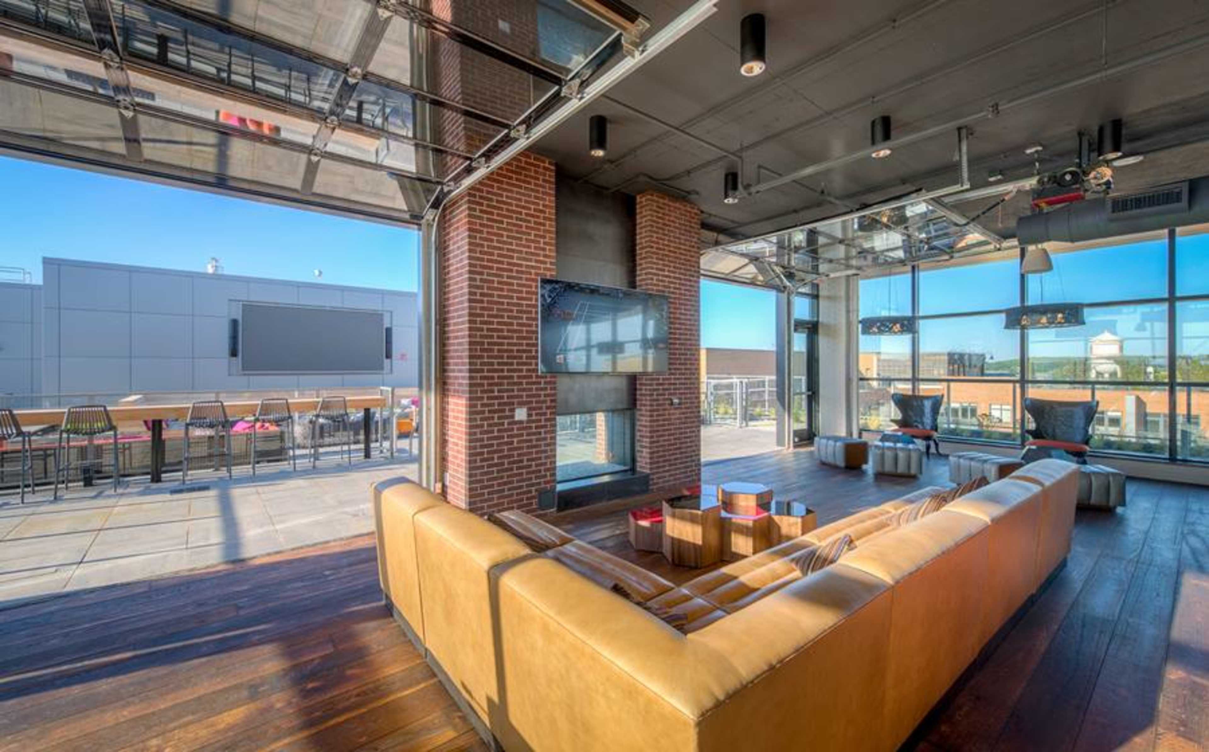 A modern lounge area with large glass windows, a brick wall, a flat-screen TV, and a spacious outdoor terrace visible in the background.