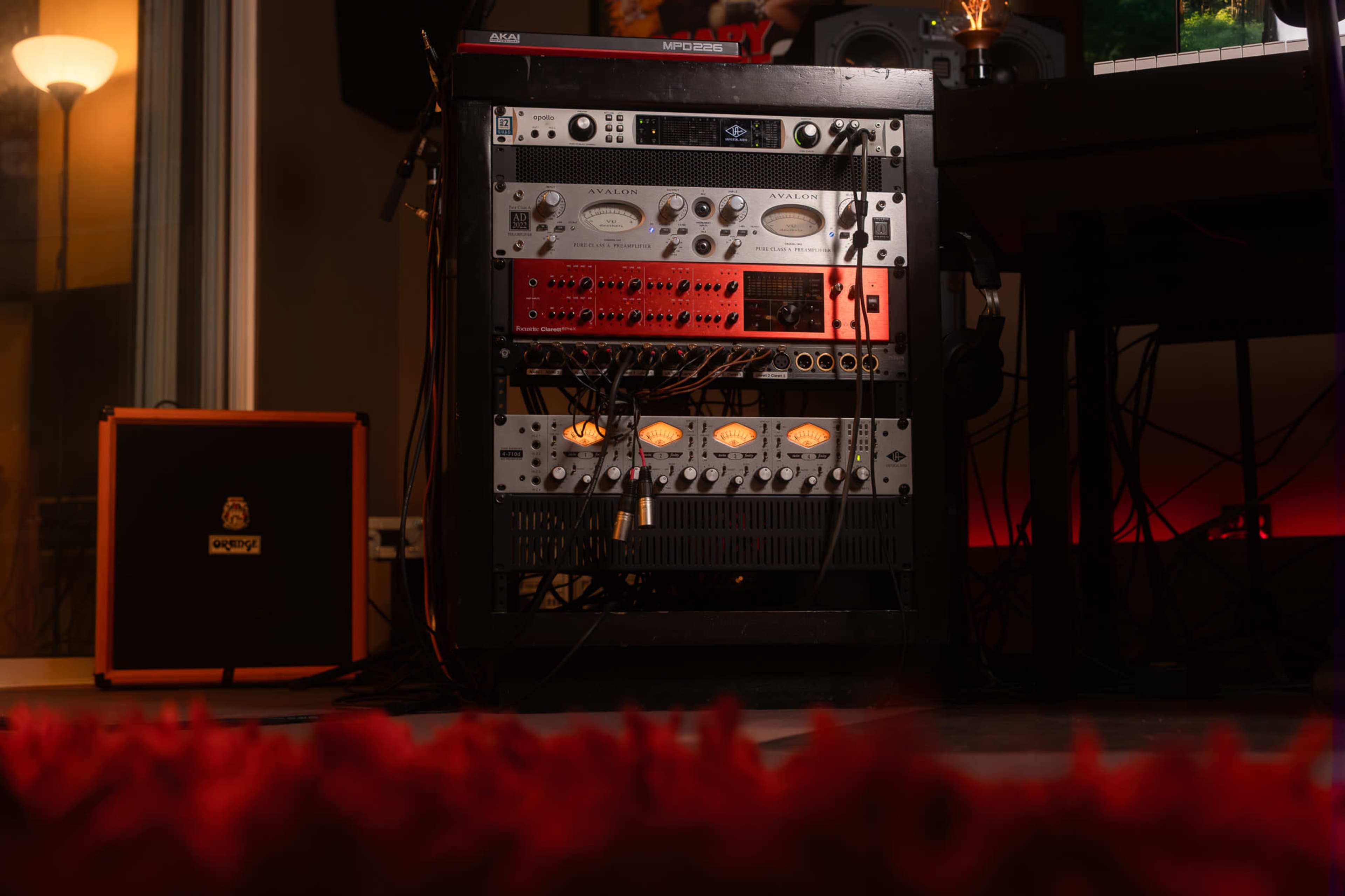 The image shows a rack of audio equipment with various processors, mixers, and connections set against a backdrop of a music studio.