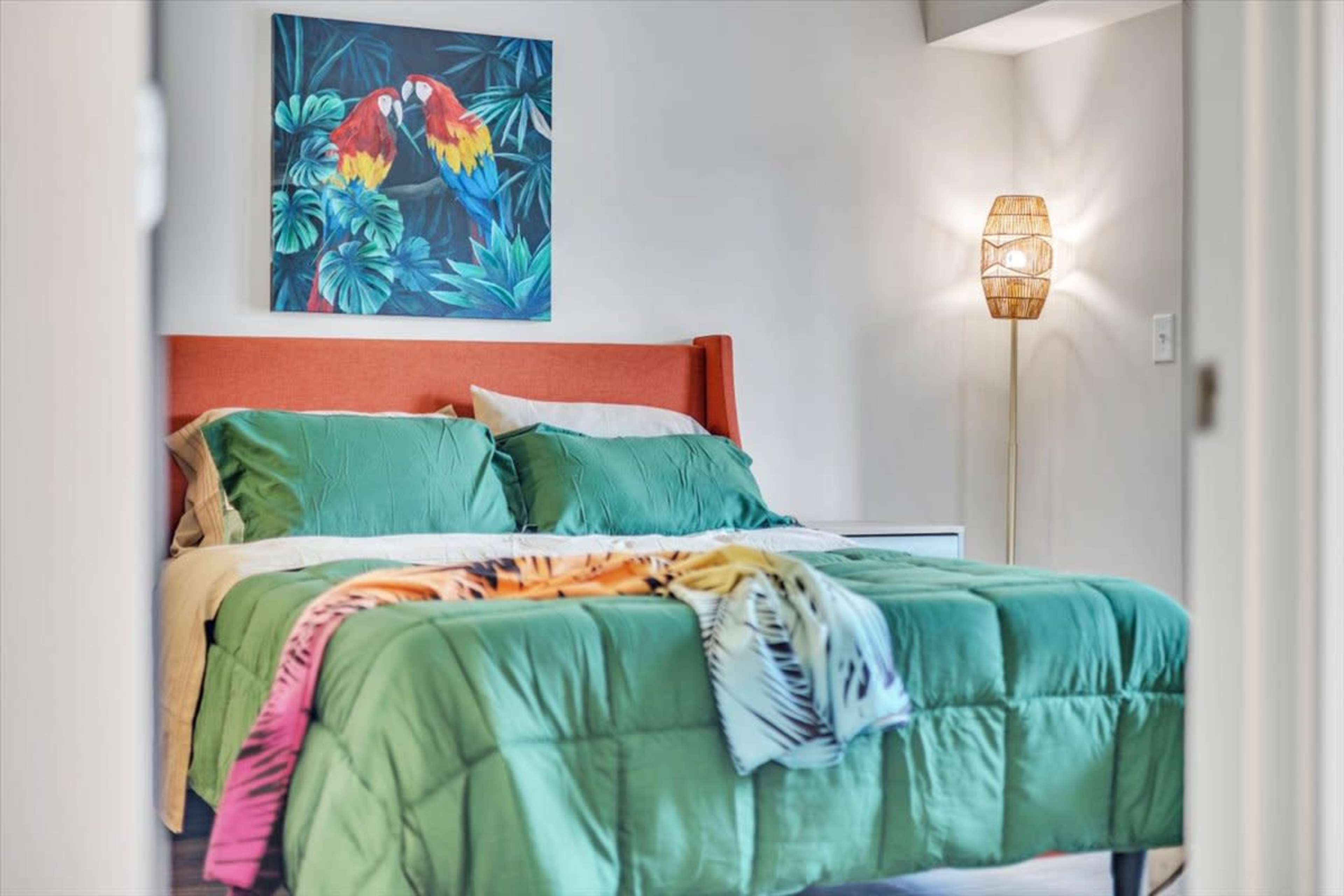 A neatly arranged bedroom features a green bedspread with a colorful throw draped on it, a painting of parrots on the wall, and a lamp beside the bed.