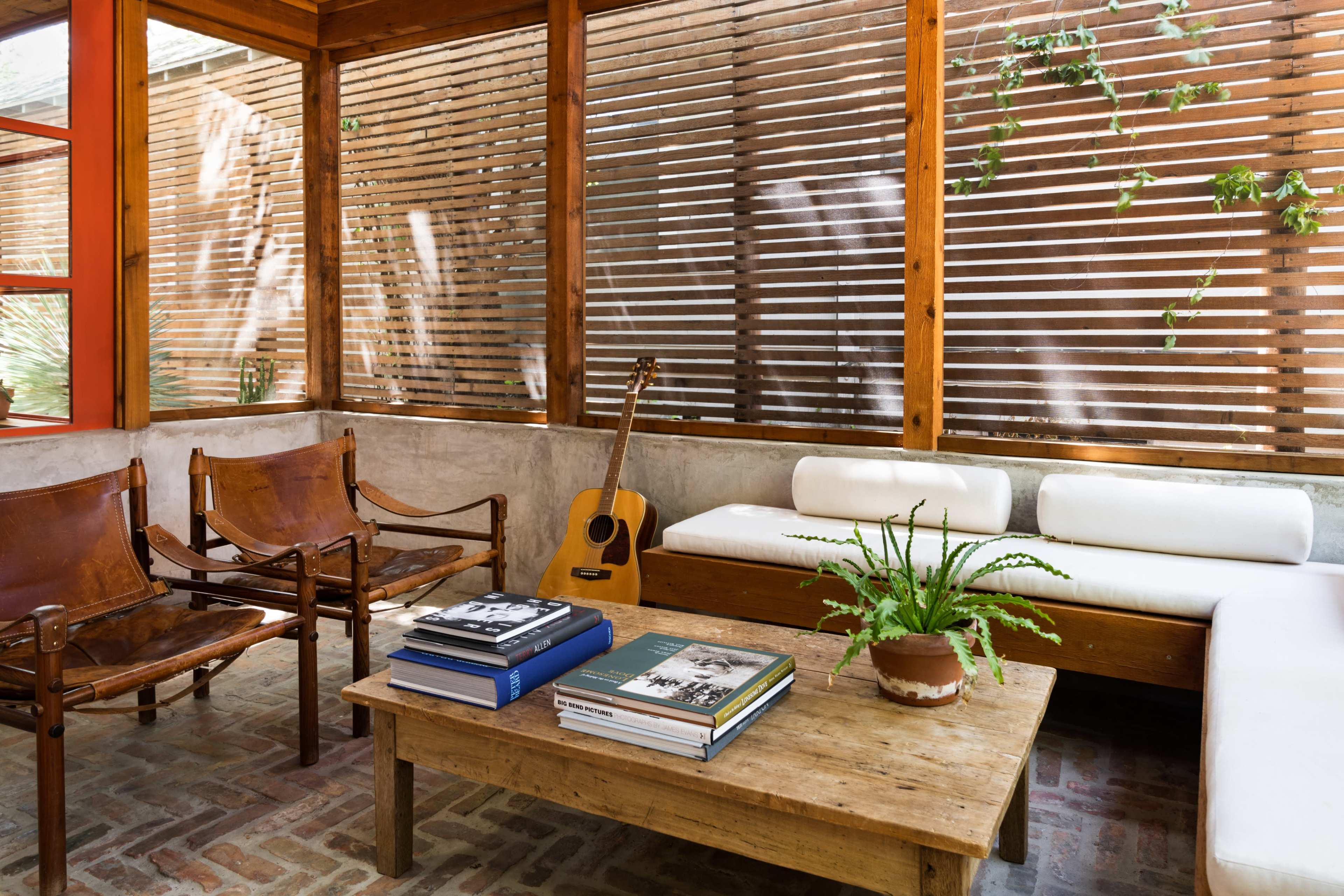 A sunlit seating area features wooden chairs and a guitar beside a coffee table stacked with books, surrounded by slatted wooden walls and greenery.