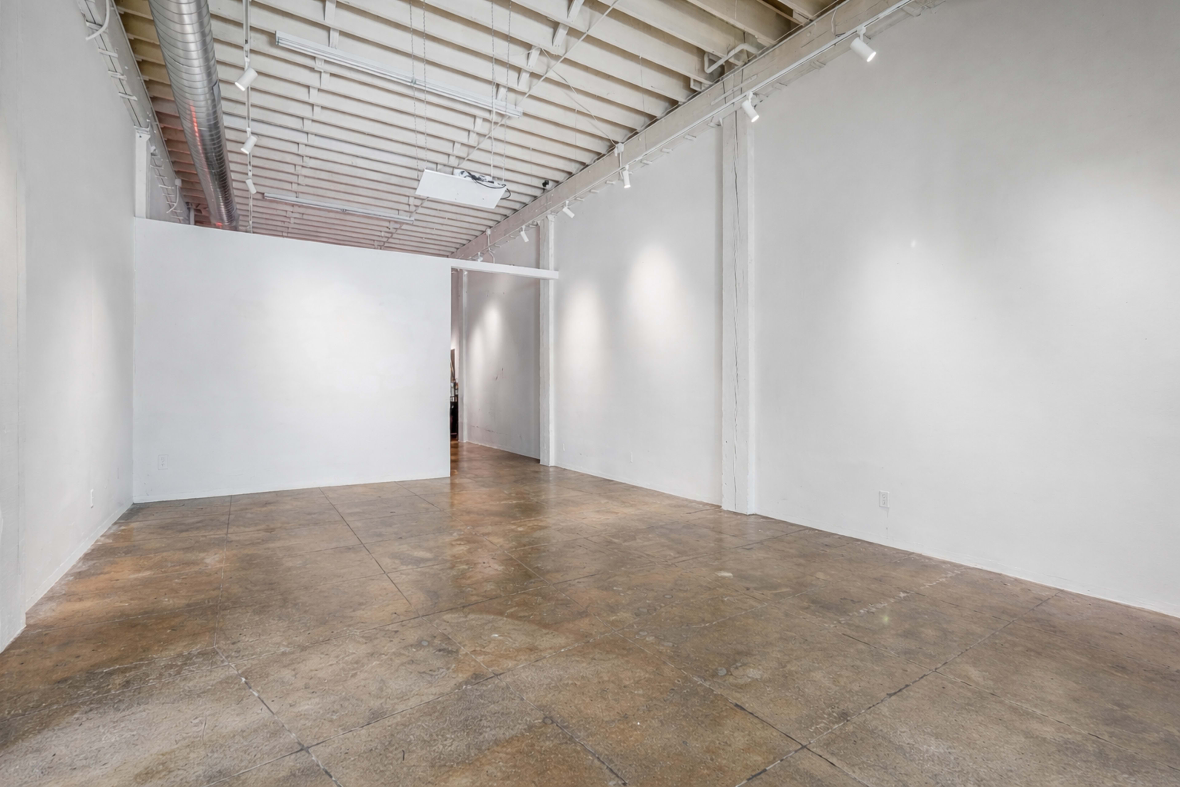 The image shows an empty, well-lit gallery space with white walls and a concrete floor.