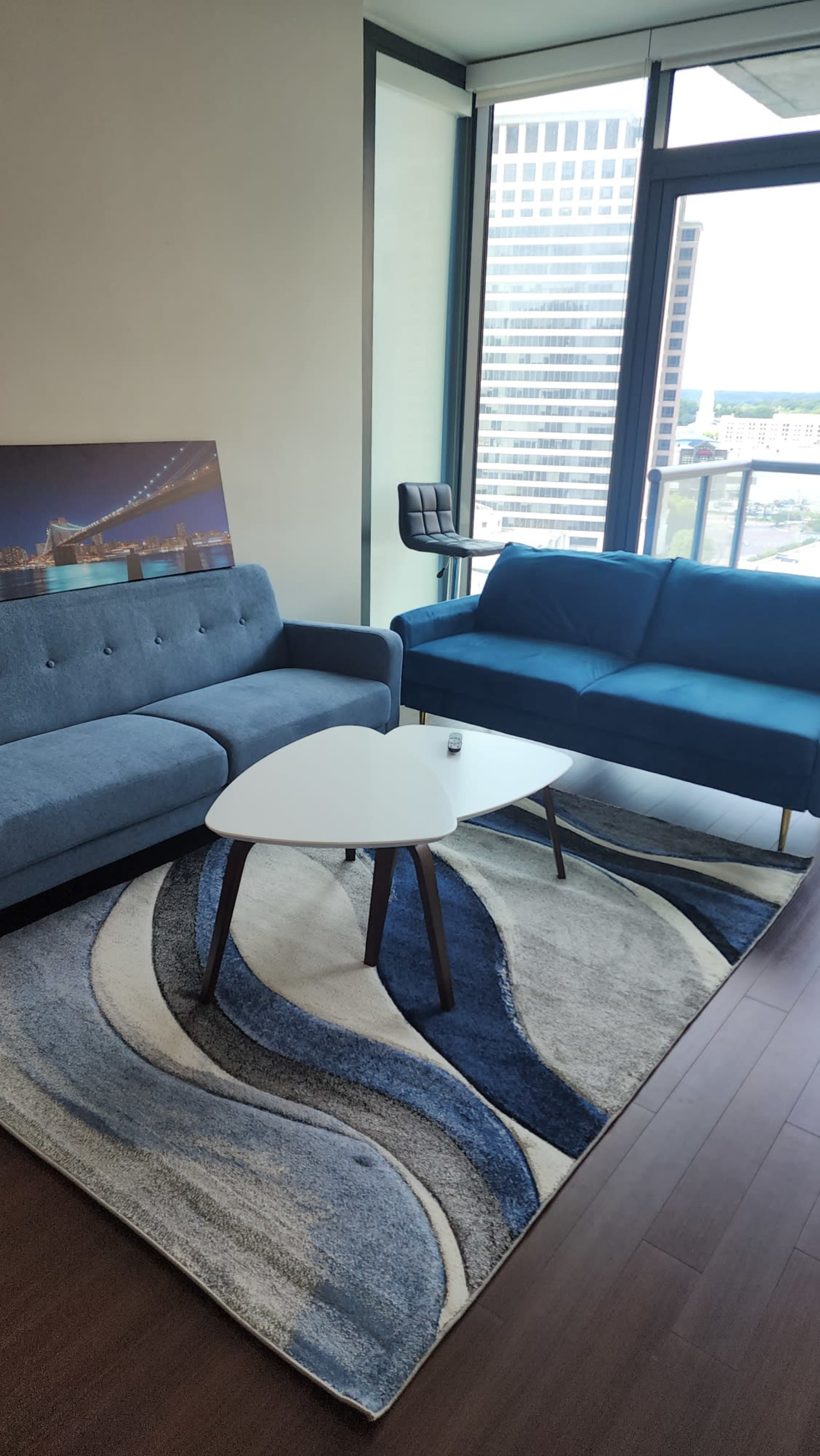 A modern living room with two blue sofas, a round white coffee table, and a large rug on wooden flooring, all set against a backdrop of large windows.